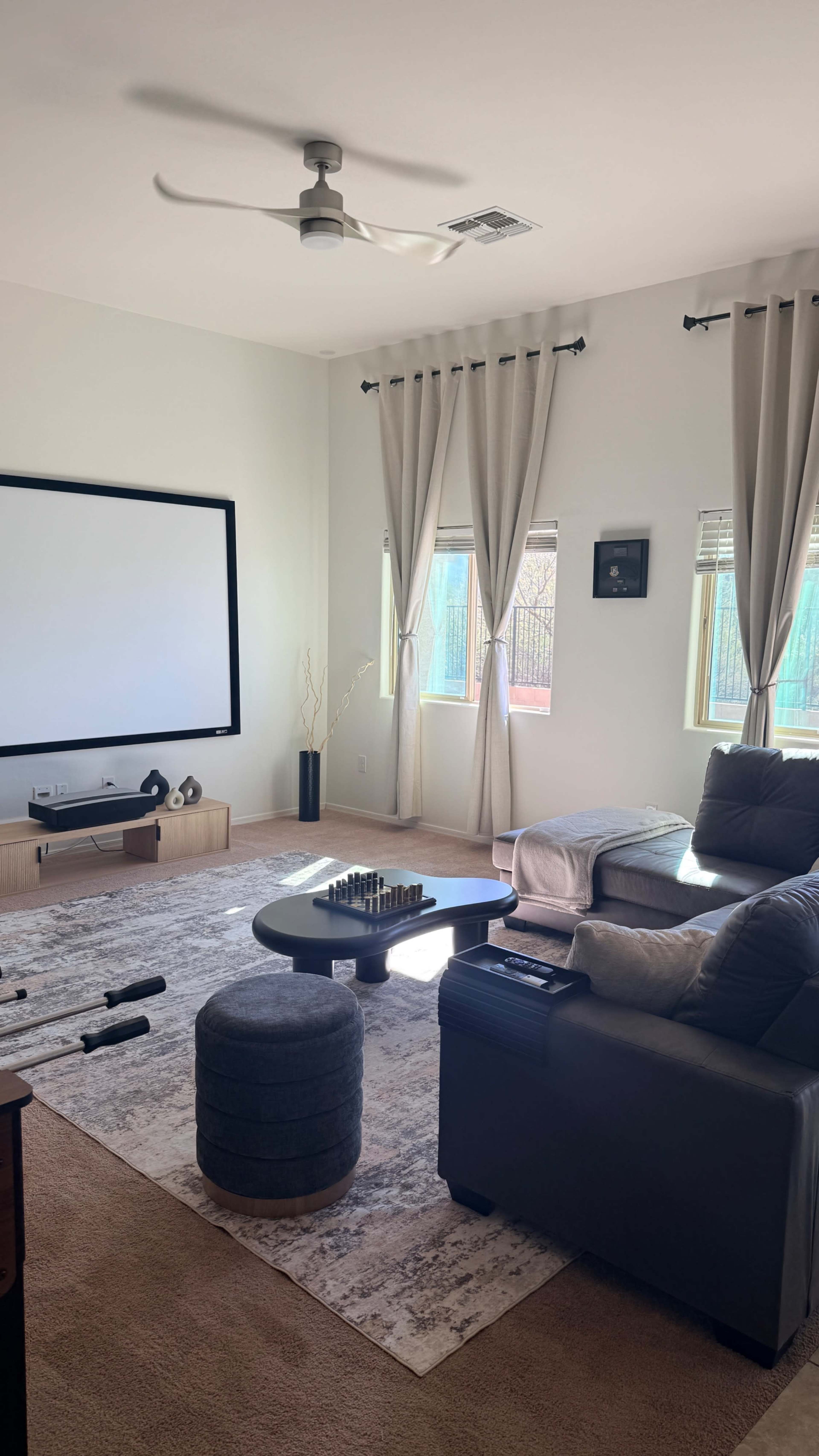 A modern living room features a large projection screen, a dark sectional sofa, a circular coffee table, and light-colored curtains.