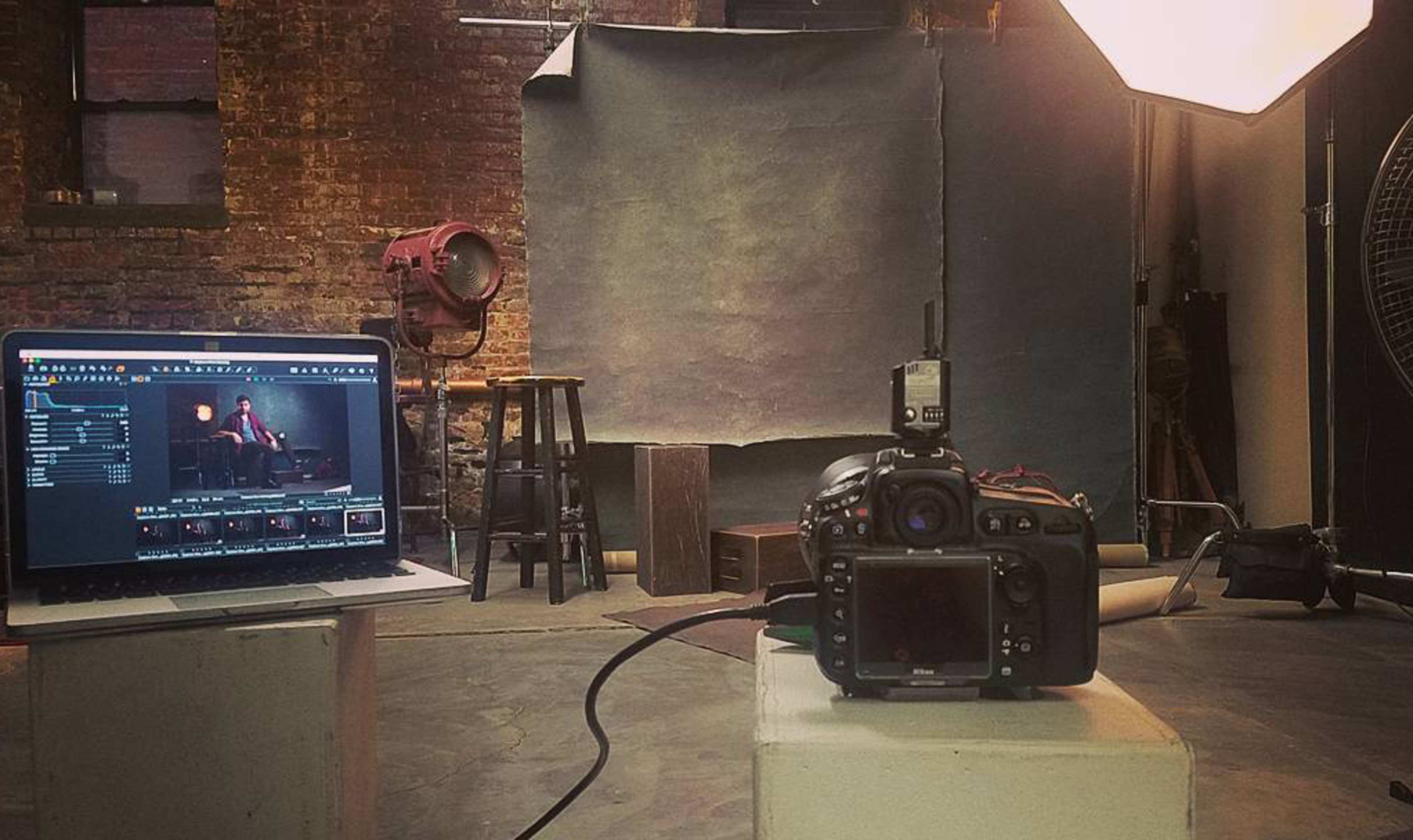 A camera is positioned on a pedestal facing a backdrop, while a laptop displays the camera's settings and images in a photography studio.