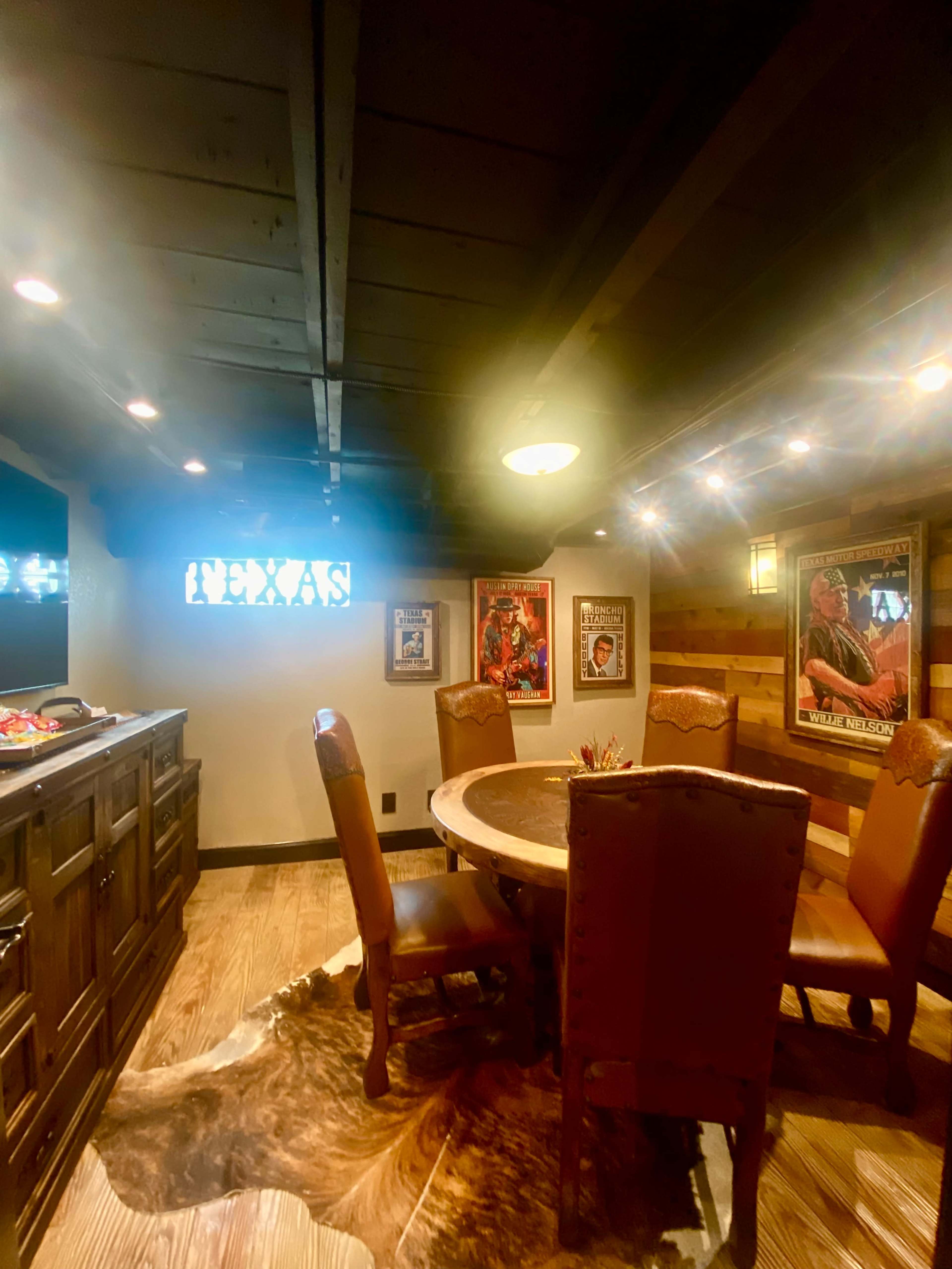 A rustic dining area features a round wooden table surrounded by upholstered chairs, with framed posters on the walls and wooden accents throughout.