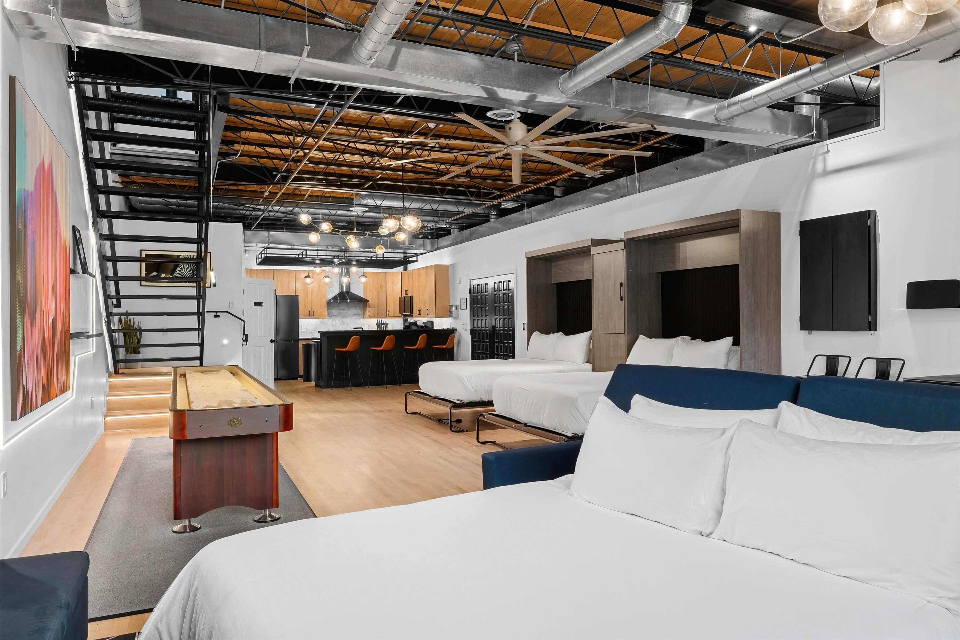 The image shows a modern open-concept living space featuring two beds, a kitchenette with seating, and a staircase leading to an upper level.