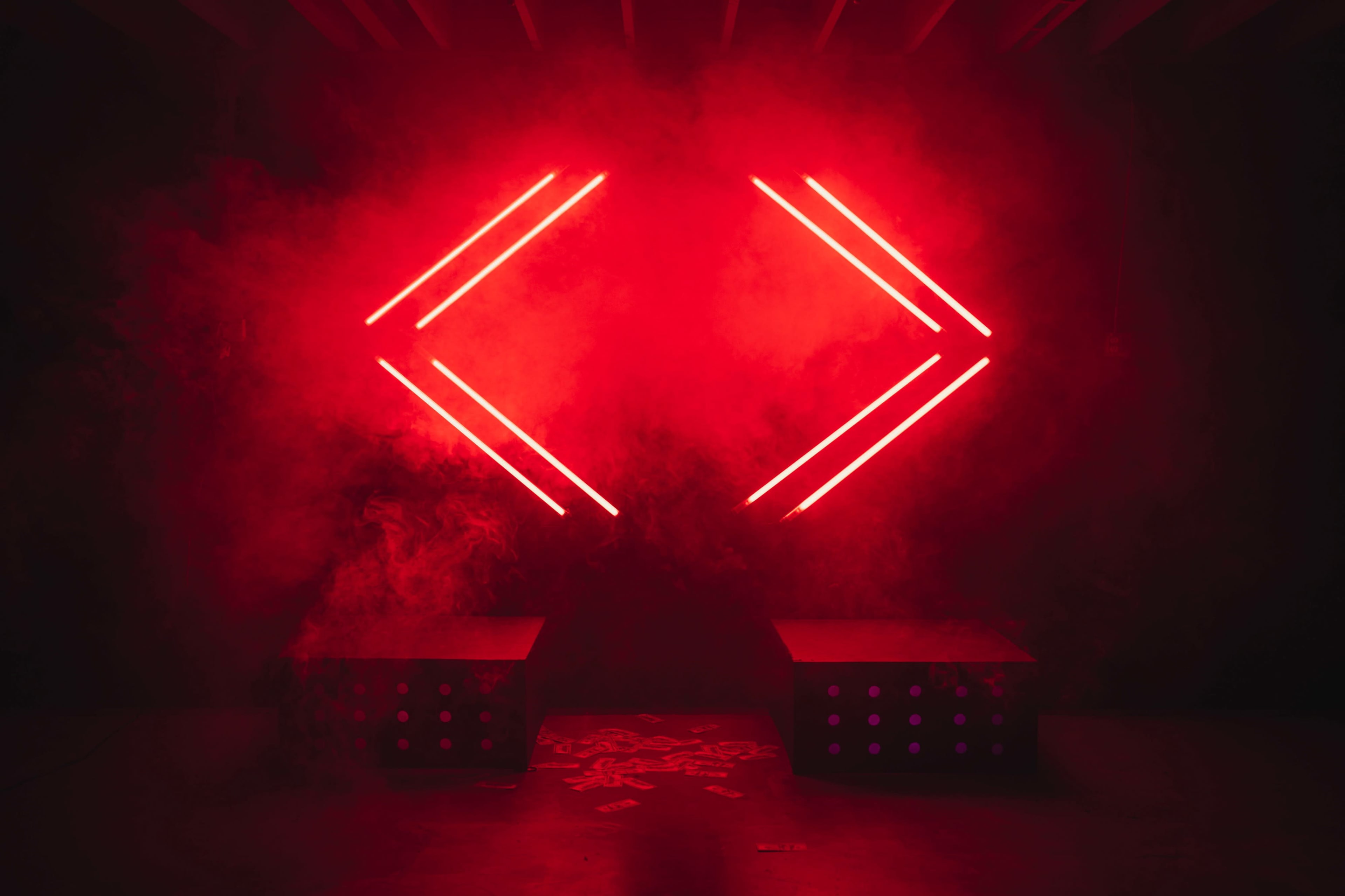 The image displays two black boxes with dotted patterns in front of a backdrop illuminated by two red neon arrows amidst a smoky atmosphere.