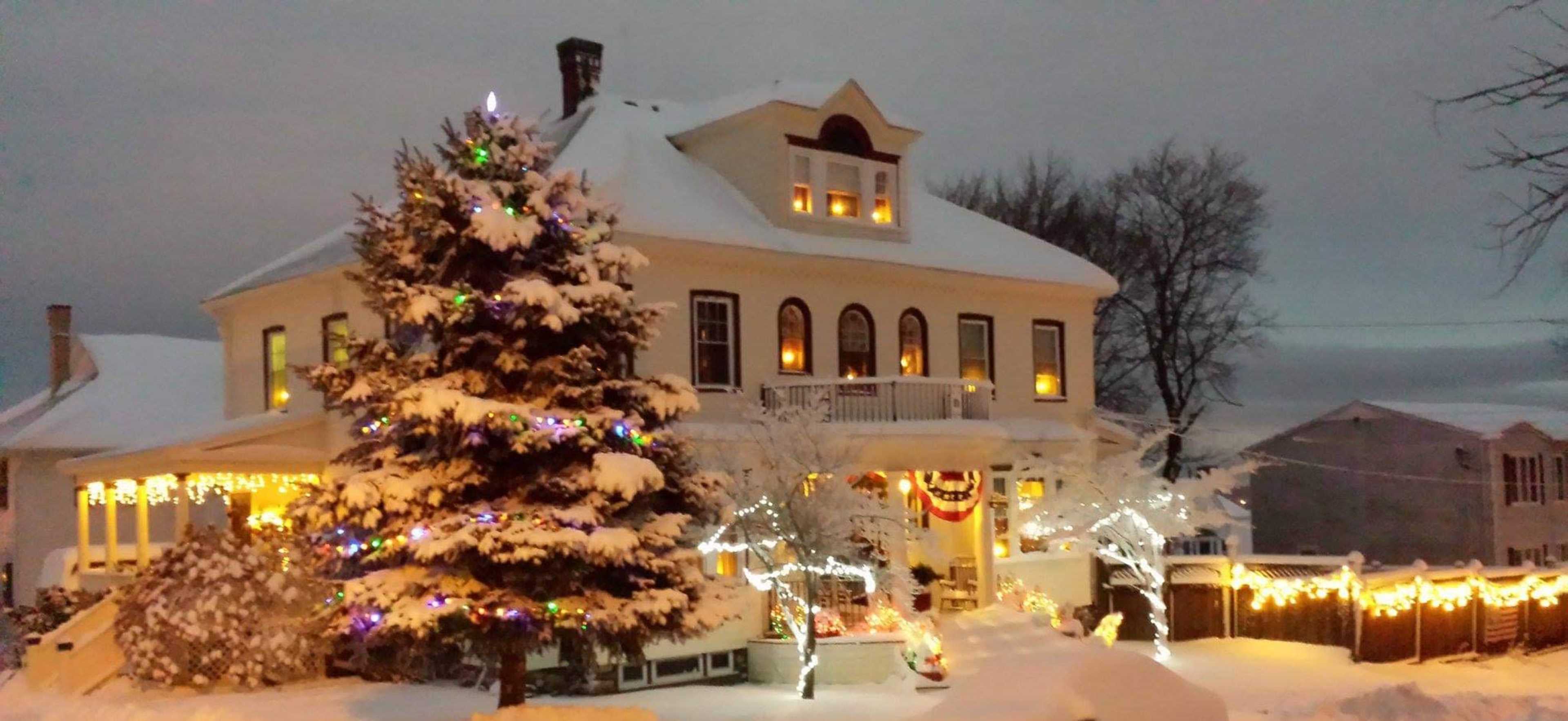A two-story house with numerous windows illuminated, surrounded by snow and decorated with colorful holiday lights and a snow-covered tree in the foreground.