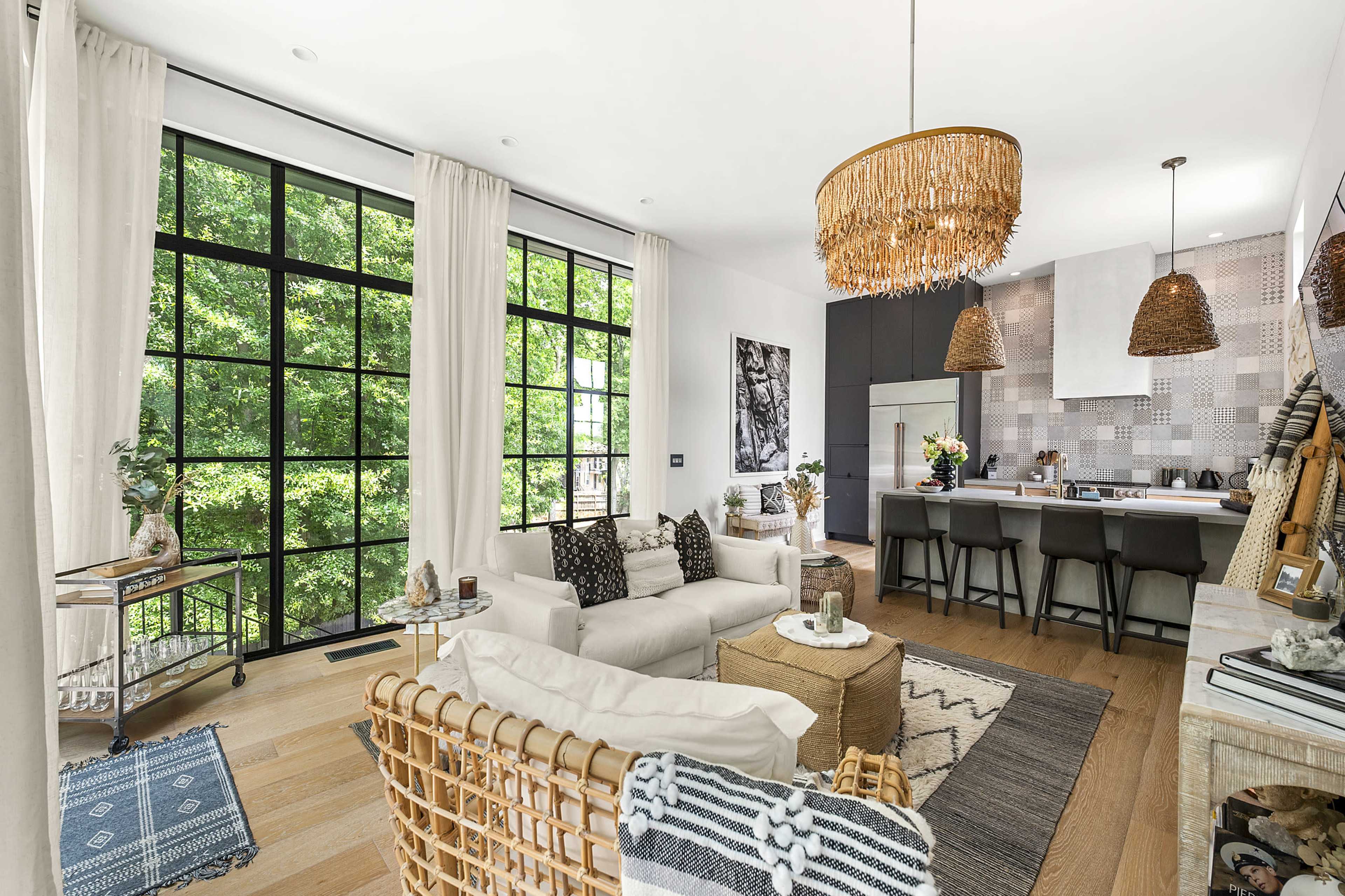 The image depicts a modern living space featuring a comfortable seating area, a kitchen with bar stools, and large windows that allow natural light and views of greenery.