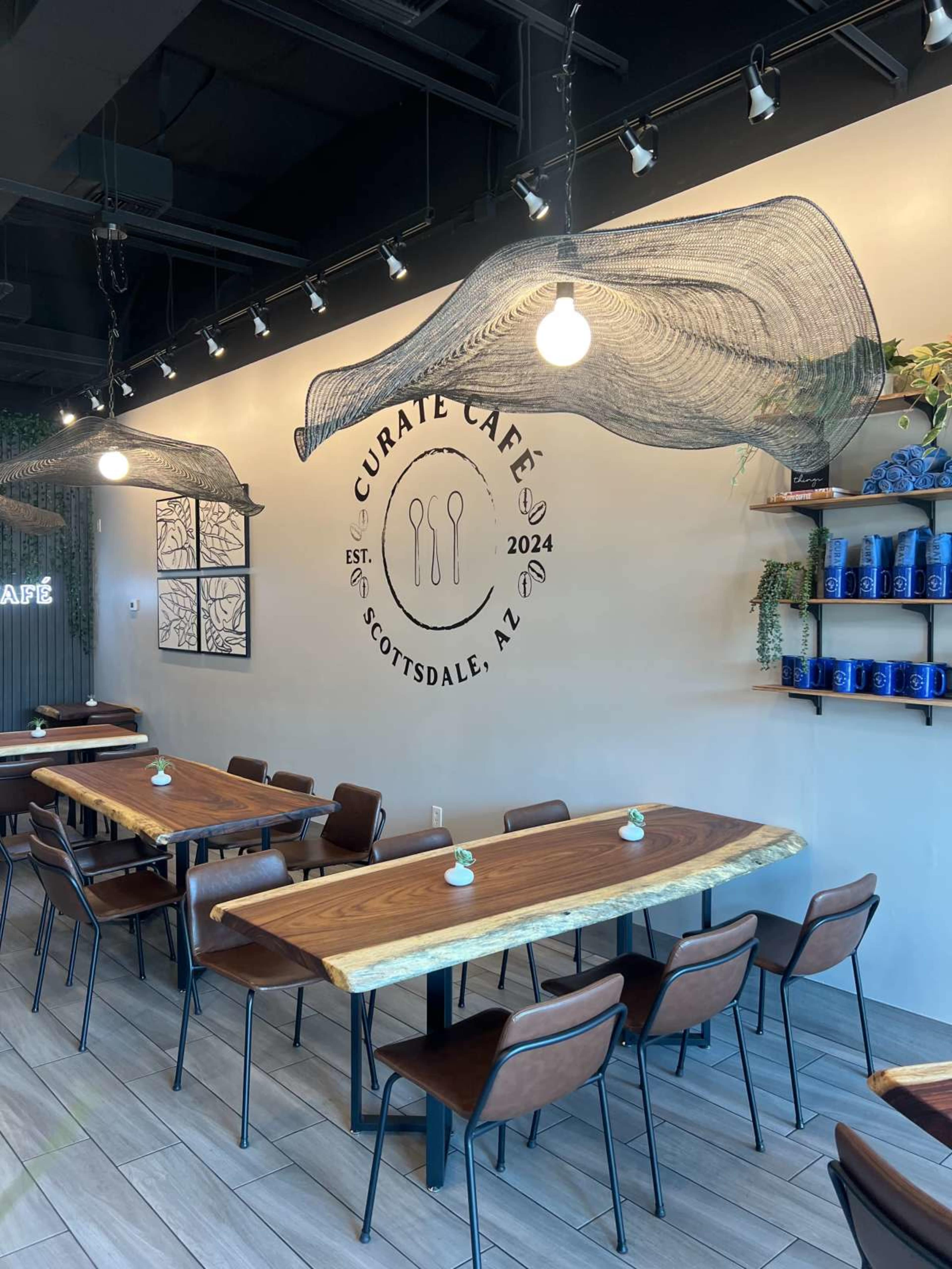 The interior of Curate Café in Scottsdale, AZ features wooden tables and a wall with decorative netting and the café's logo.
