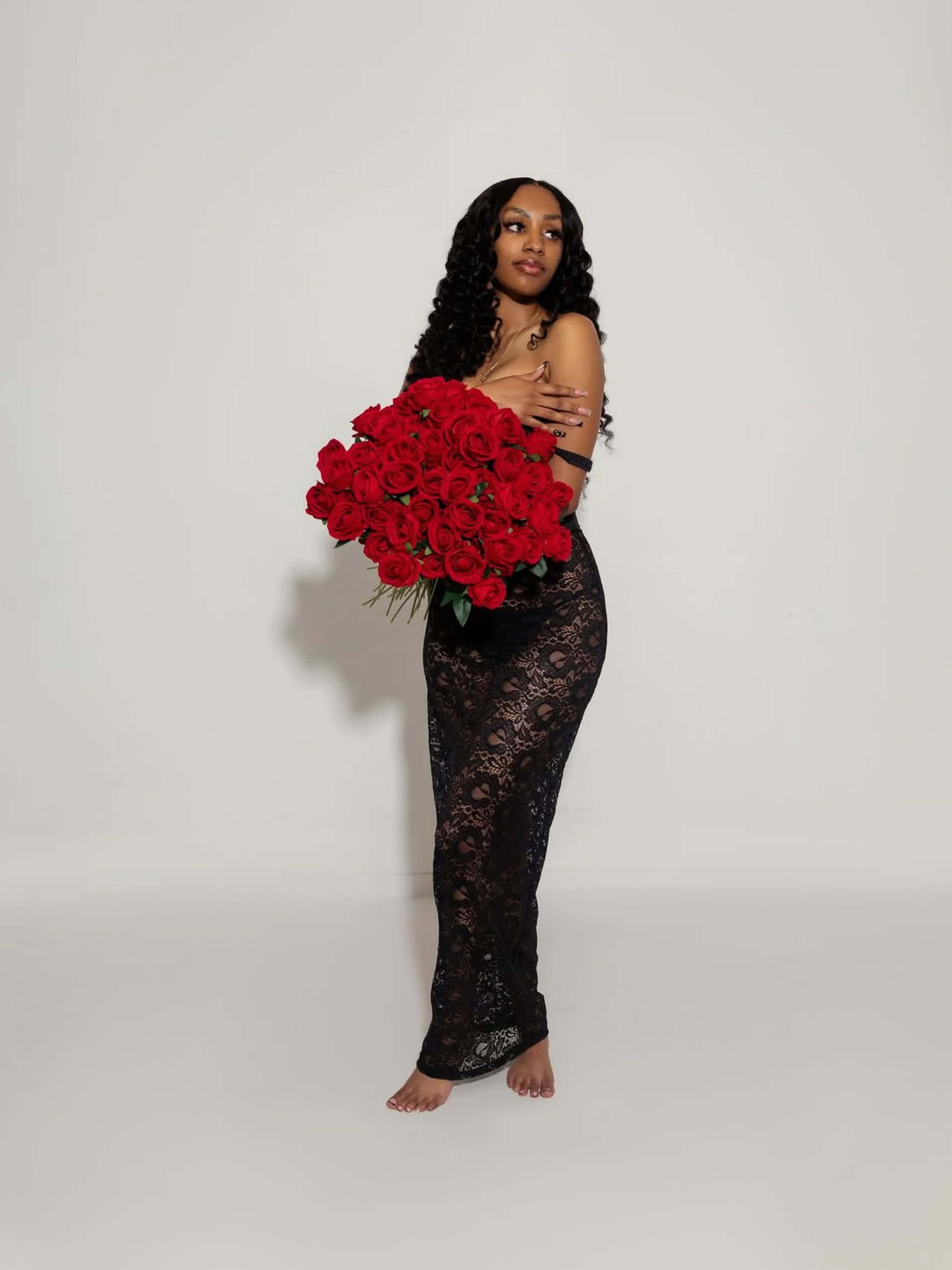 A woman in a black lace dress holds a large bouquet of red roses while standing on a plain white background.