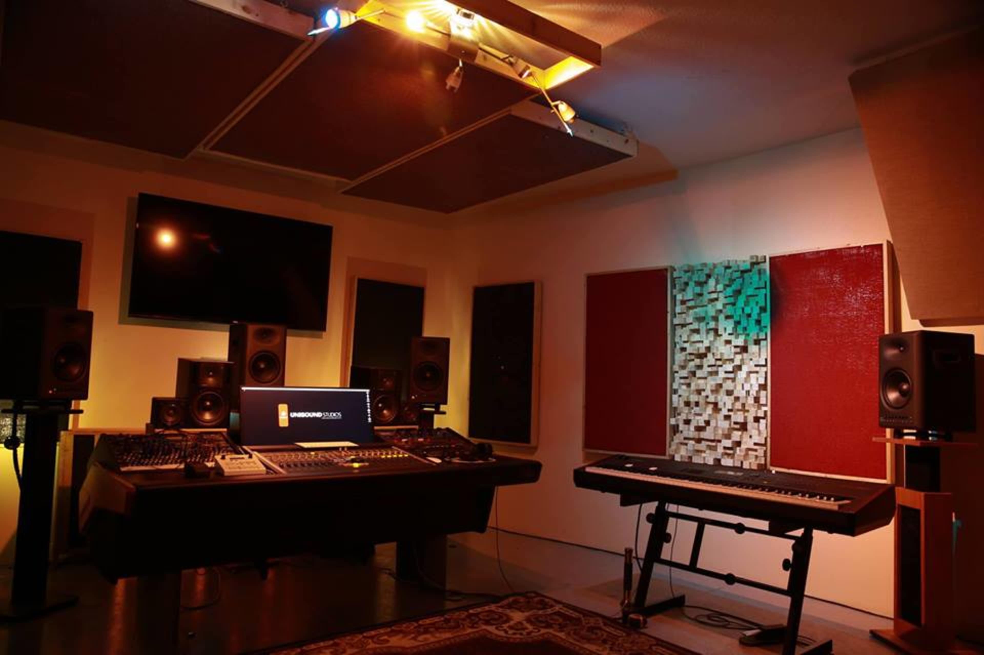 A recording studio featuring a mixing console, studio monitors, and a keyboard with soundproof panels on the walls.