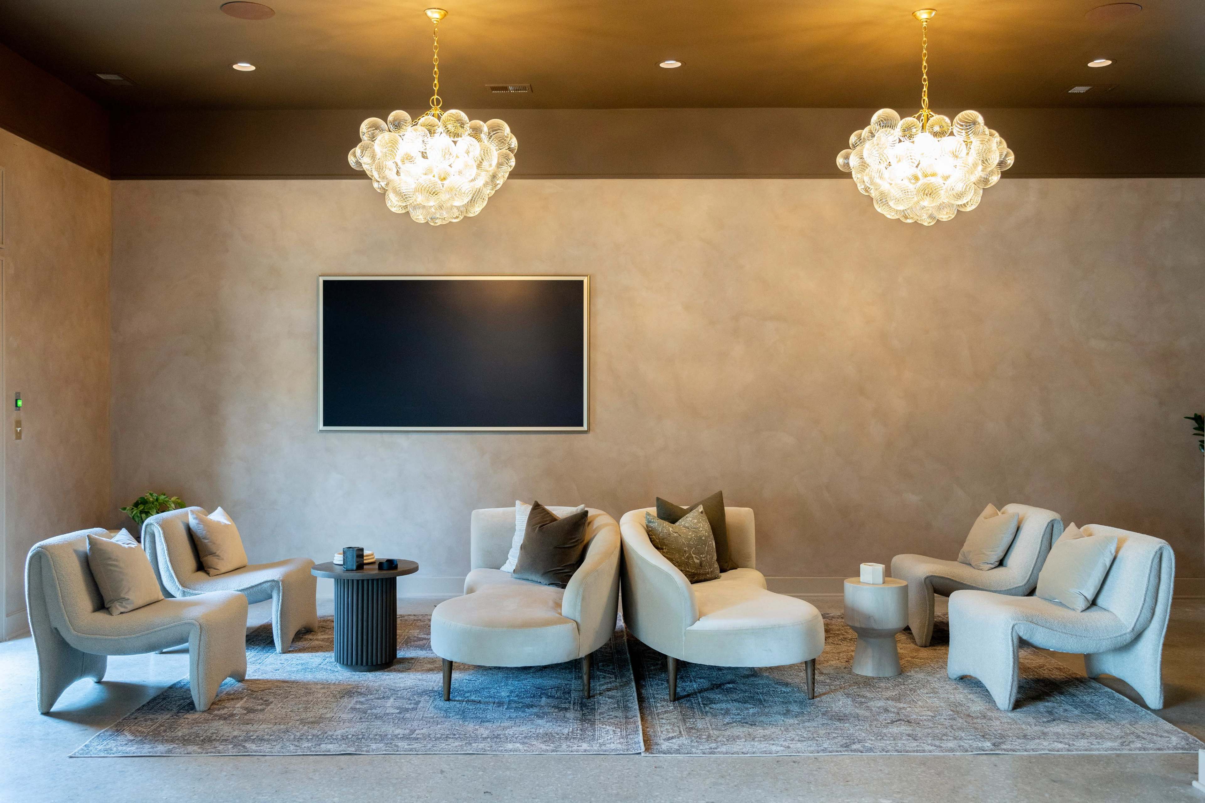 A modern lounge area featuring light-colored furniture arranged around a central table, with two decorative chandeliers hanging above.