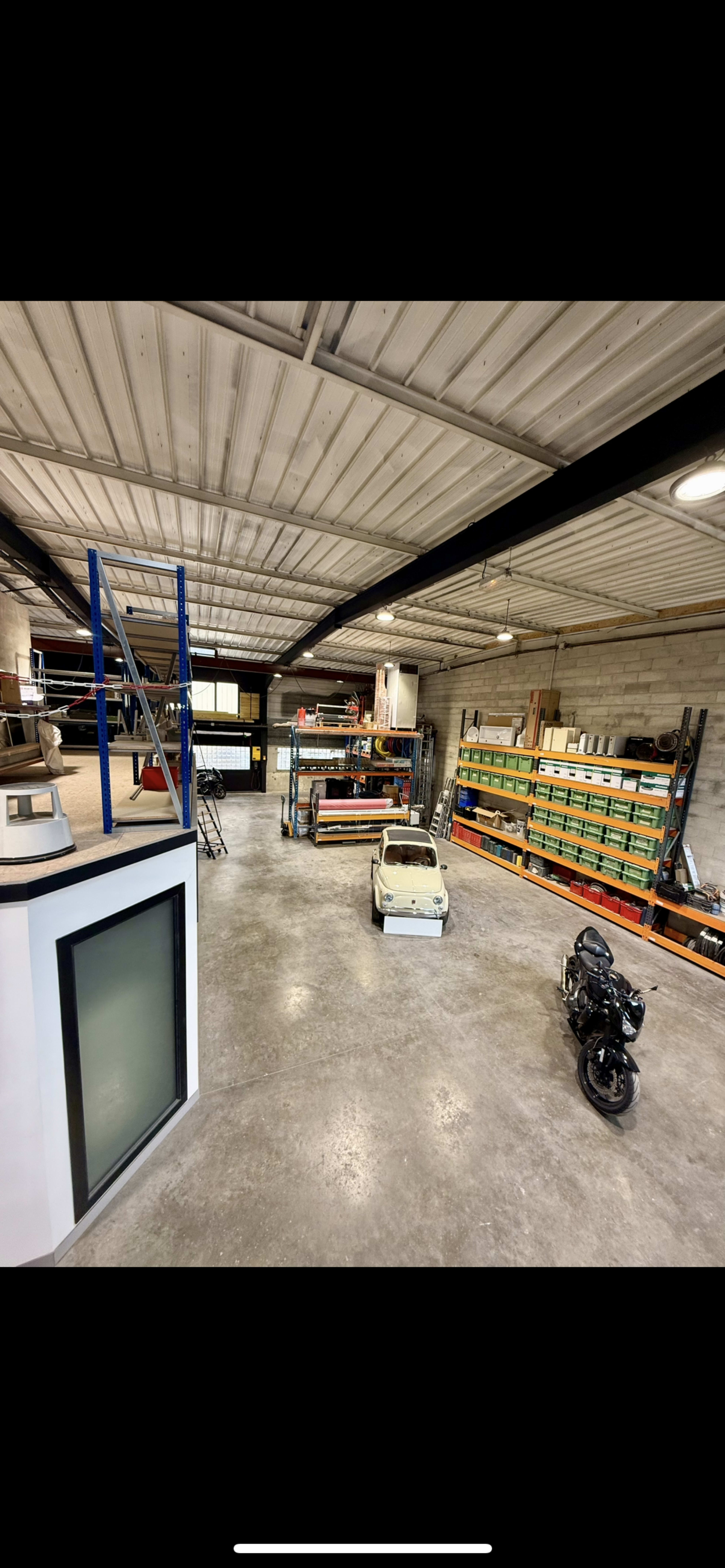 The image shows a spacious industrial garage with shelving units filled with stored items, a motorcycle parked on the floor, and a classic car positioned in the center.