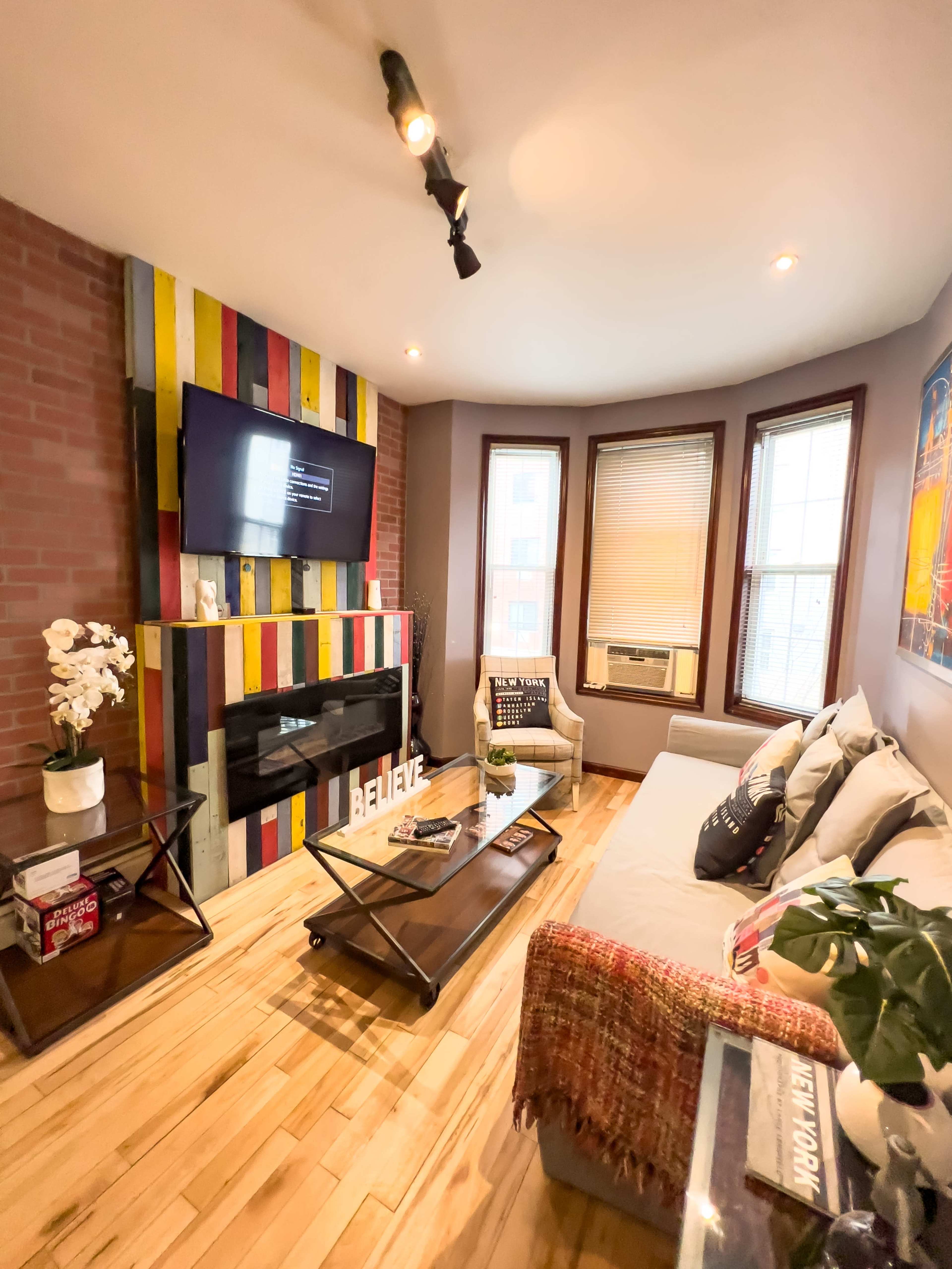 A cozy living room features a modern couch, a colorful accent wall with a mounted television, and hardwood floors, illuminated by natural light from bay windows.