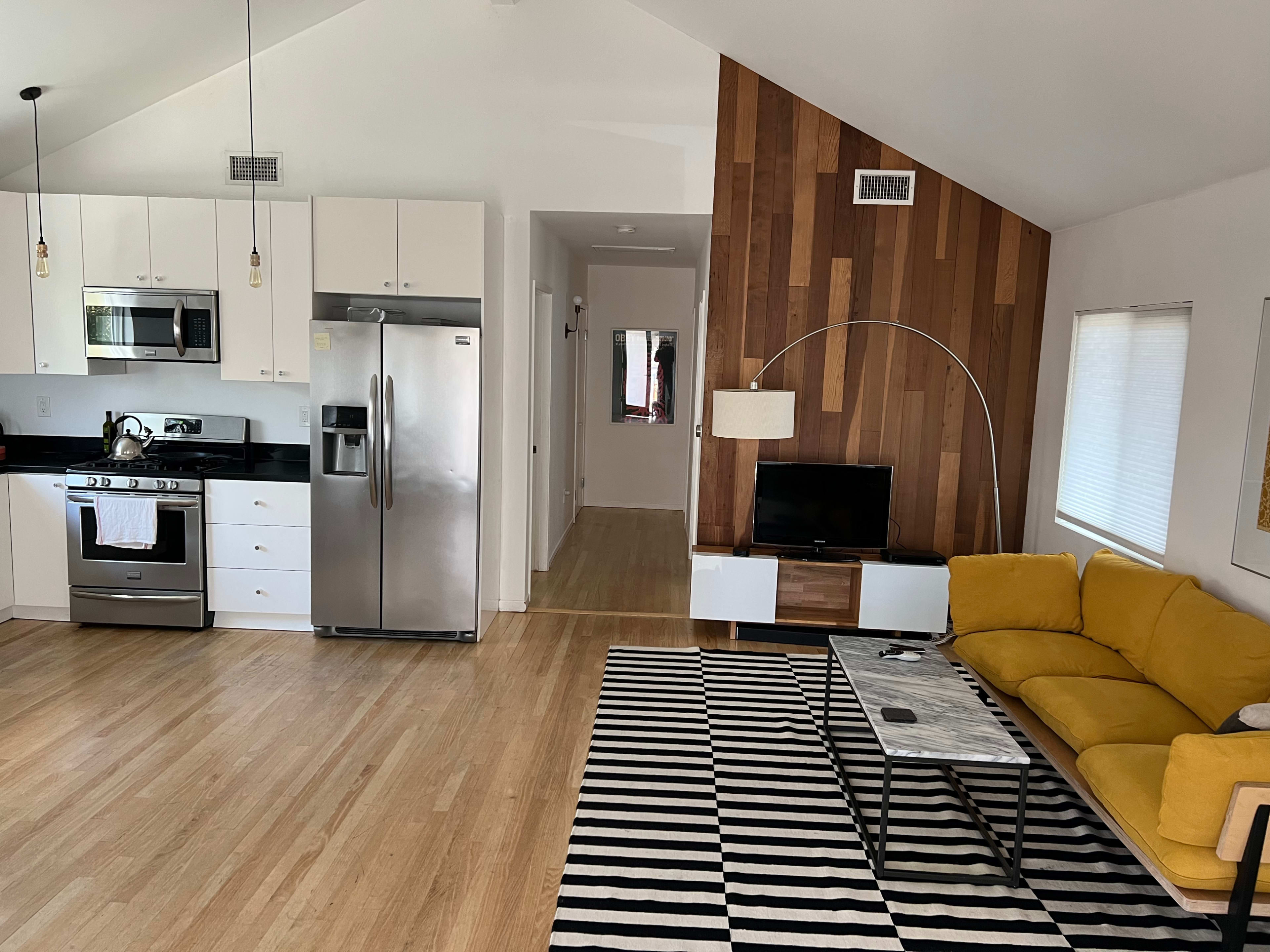 The image shows a modern open-concept living area with a kitchen, a yellow sofa, a striped rug, and a wood-paneled accent wall.