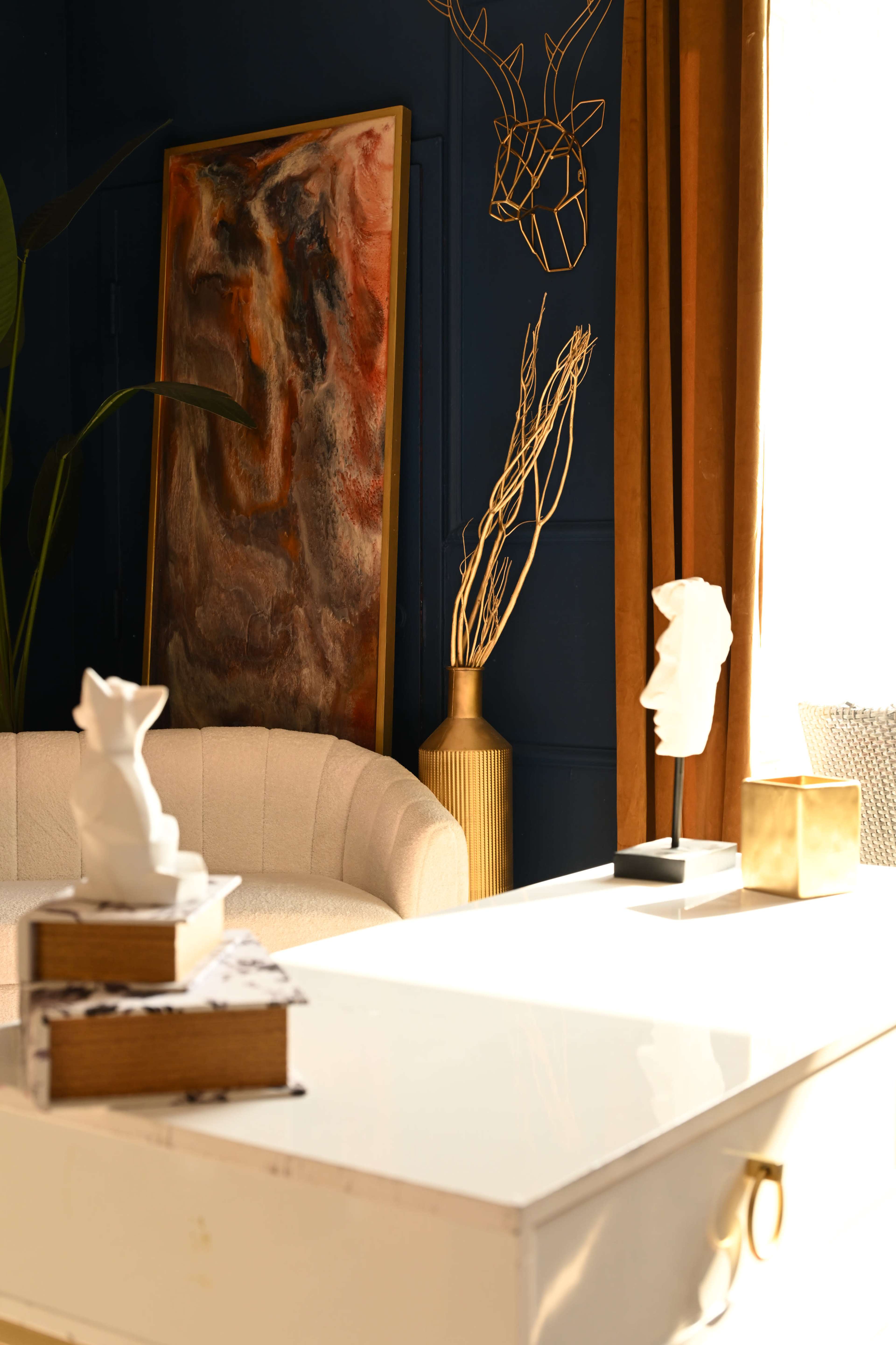 The image shows a modern living room with a cream-colored sofa, a gold-accented sculpture, and a colorful abstract painting on a dark blue wall.