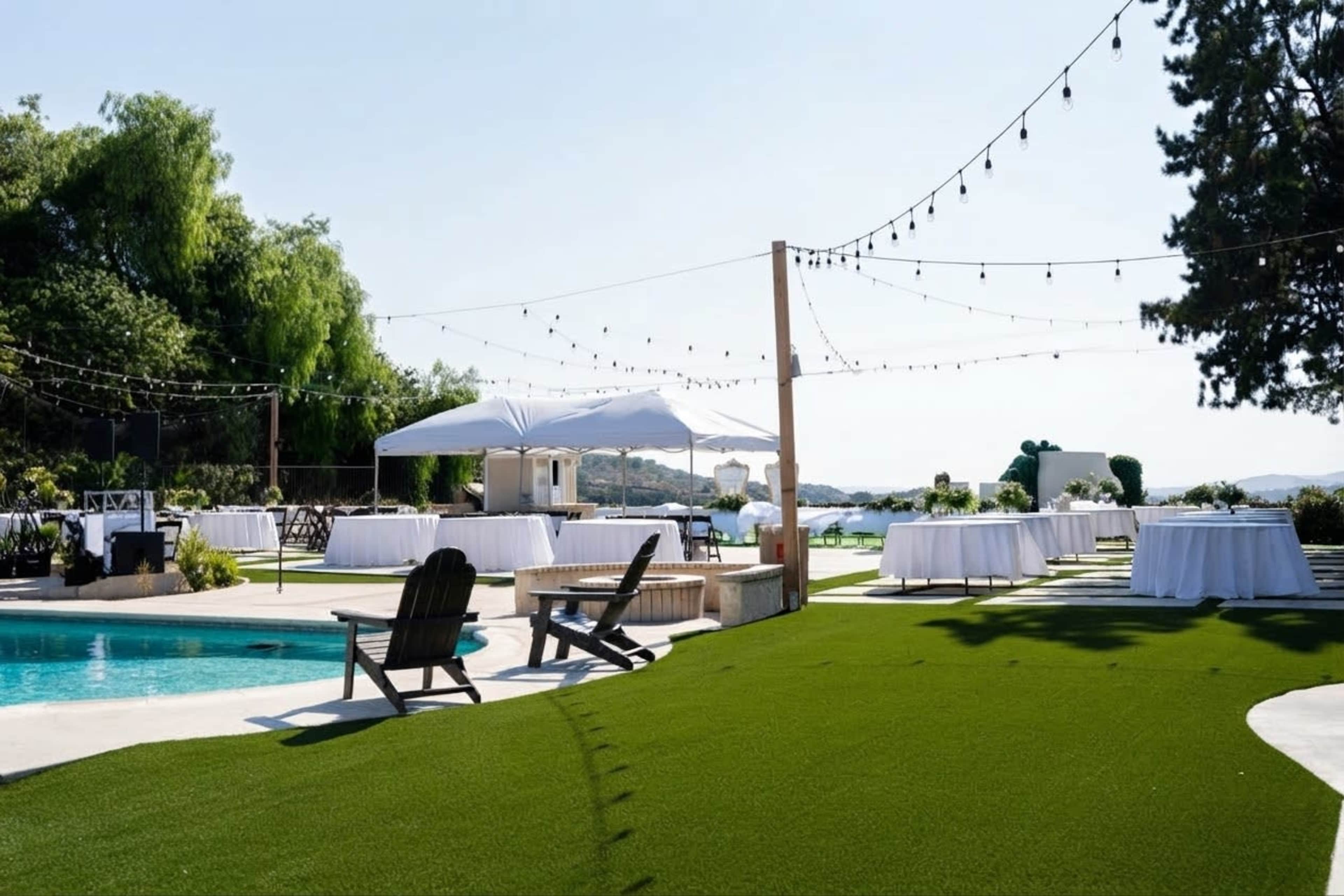 The scene features an outdoor venue by a pool with set-up tables under white tents and string lights overhead, surrounded by green grass and a view of the water.