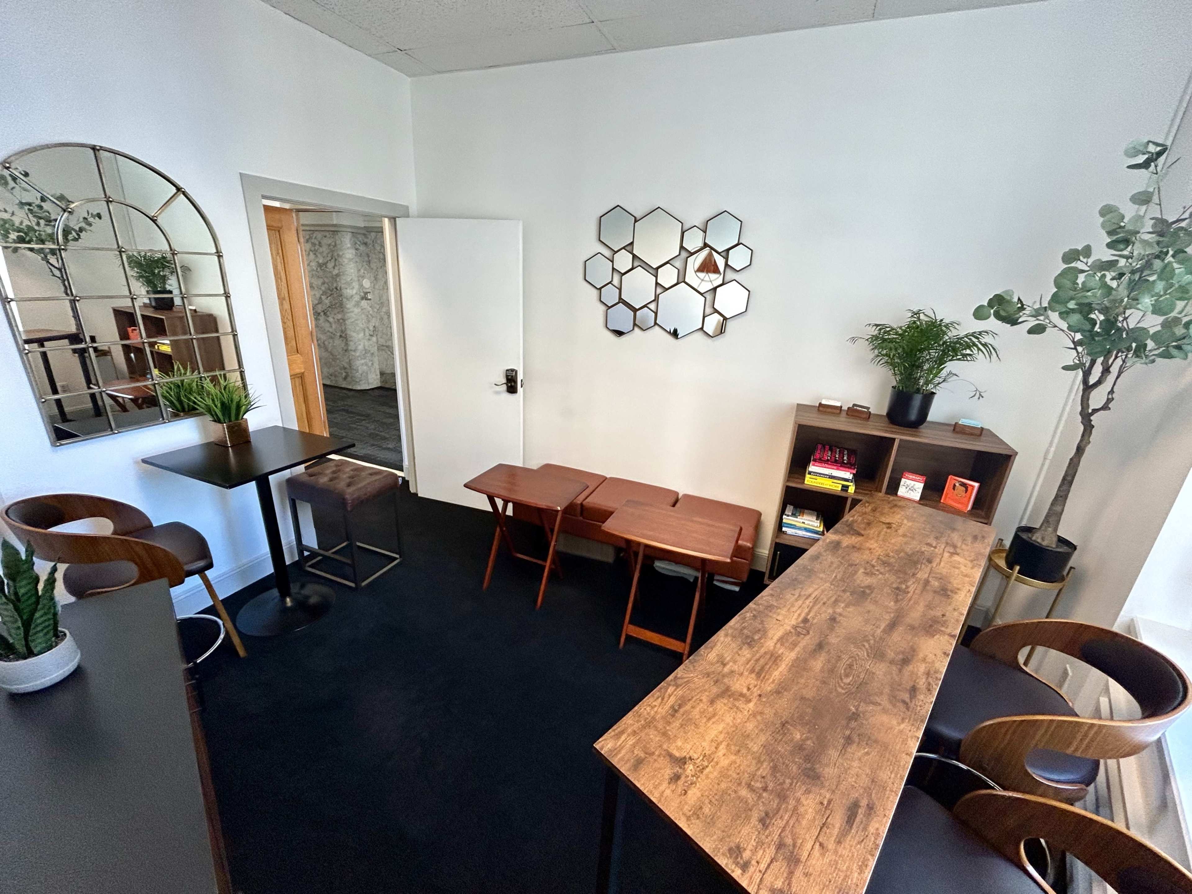 A minimalist interior features a wooden table, three stools, and a bookcase against a white wall, complemented by plants and decorative mirrors.
