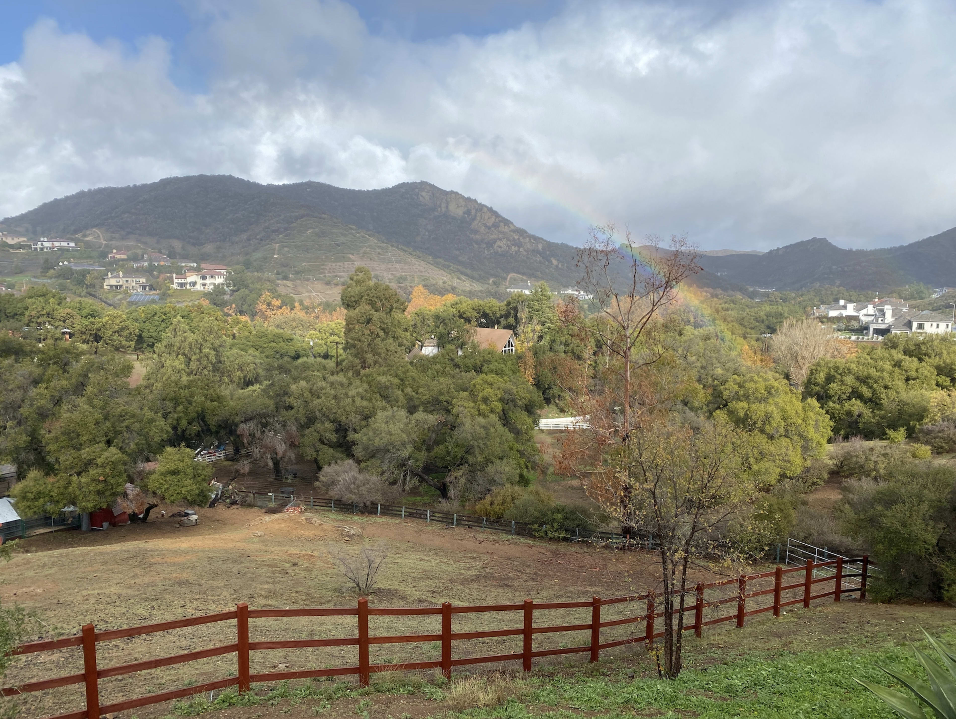 A wooden fence borders a grassy field with scattered trees and a rainbow arching over the distant mountains.