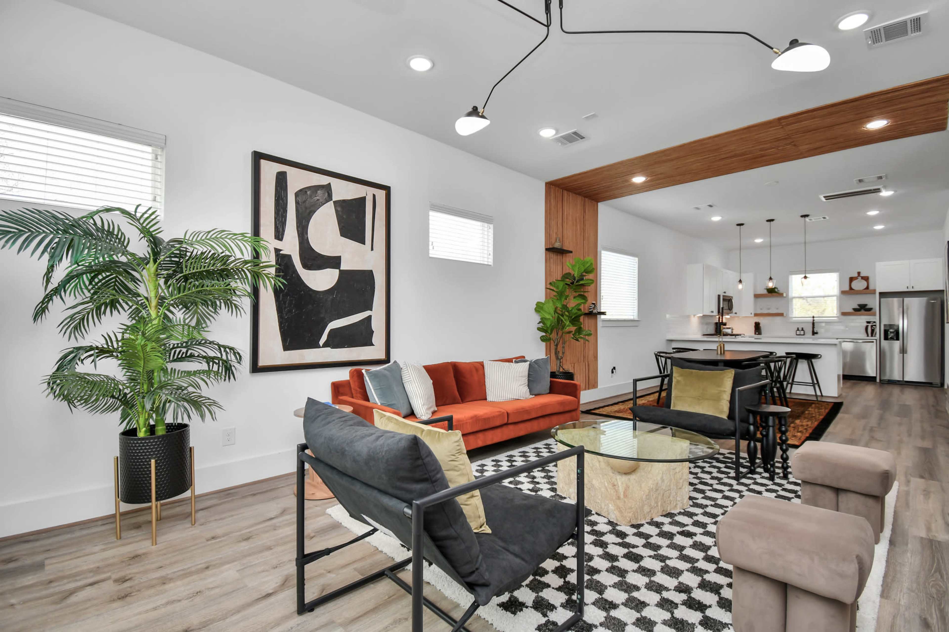 The image depicts a modern living room and kitchen area featuring a mix of seating options, a large abstract artwork on the wall, and natural elements like a potted plant.