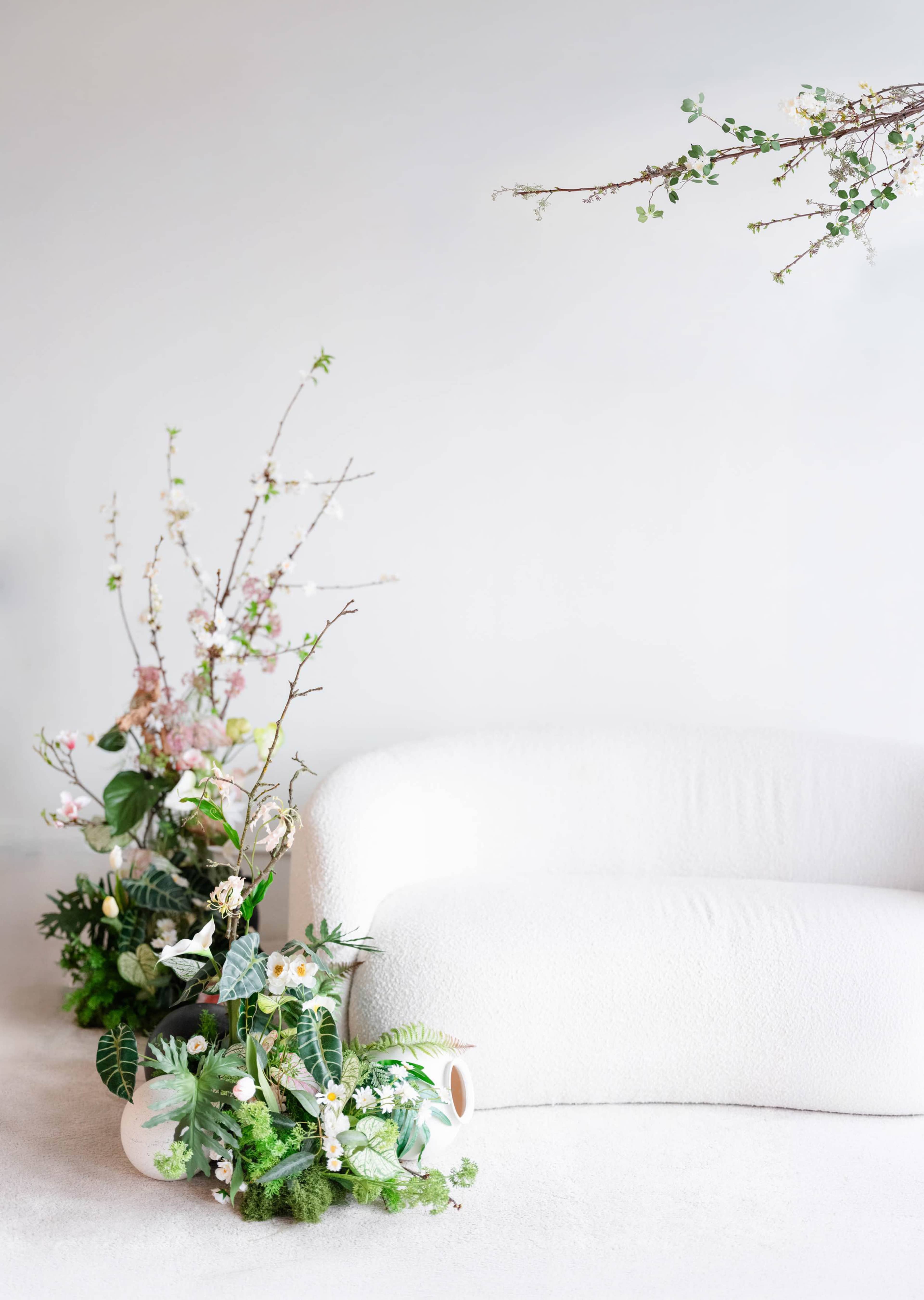 A white, textured sofa is positioned against a plain wall, surrounded by an arrangement of various green plants and flowering branches.