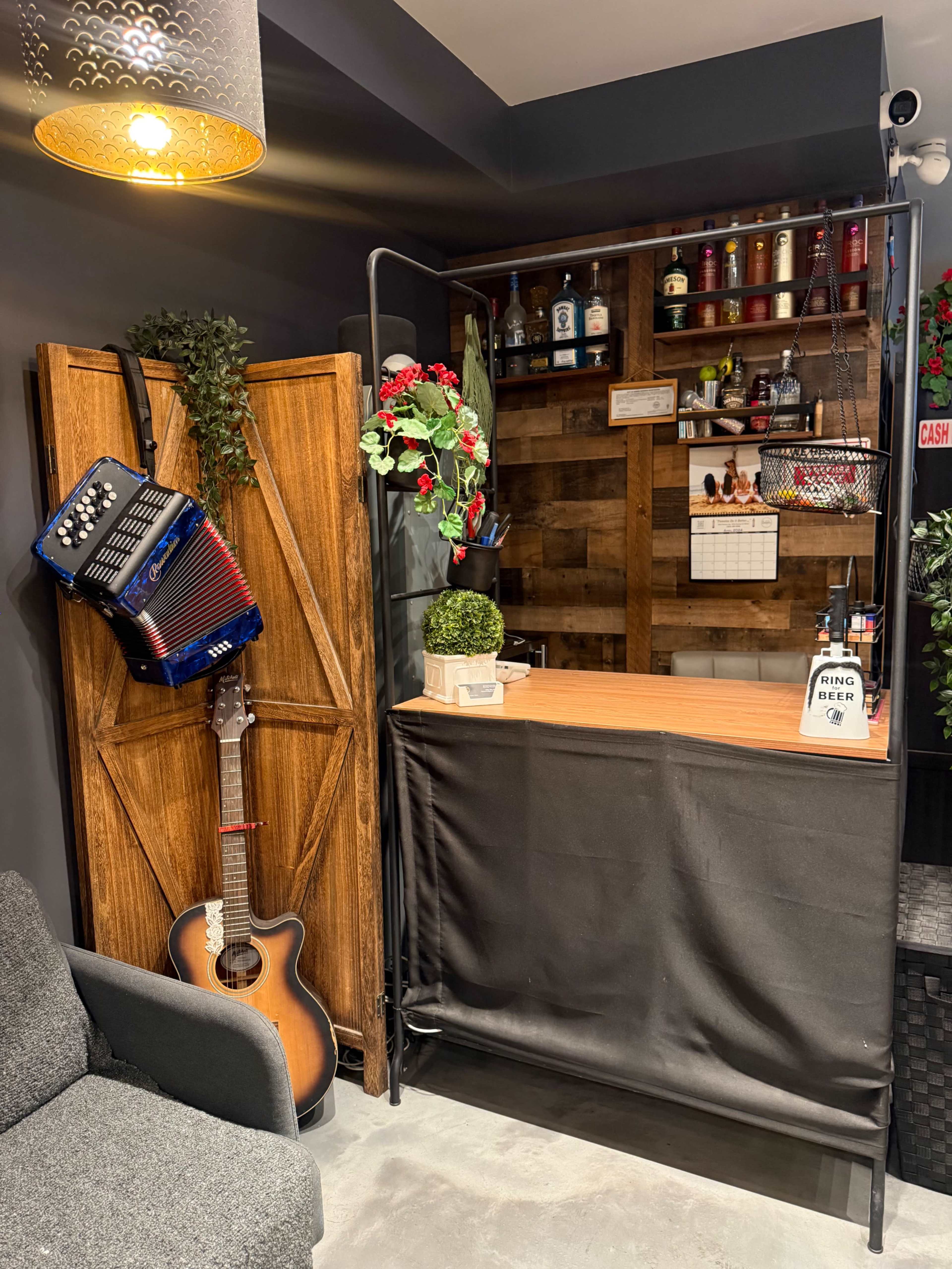 The image shows a bar counter made of wood and metal, decorated with bottles, plants, and an accordion, alongside a guitar leaning against the side.