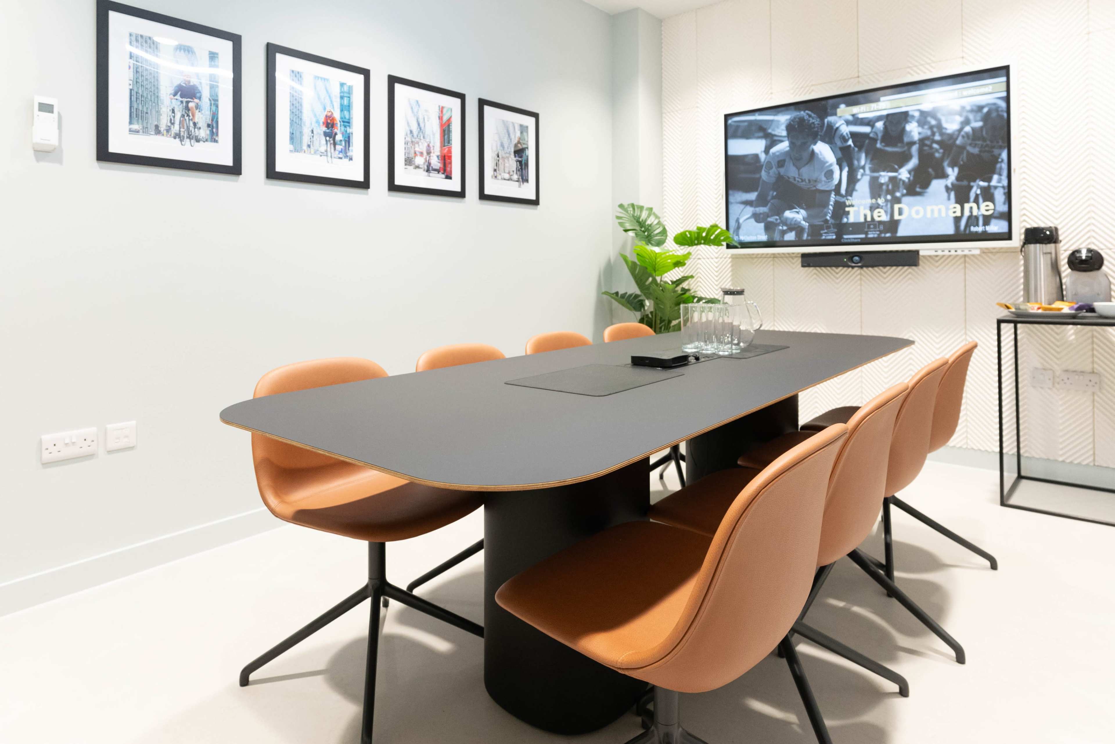 A modern conference room features a sleek black table surrounded by brown chairs, with framed artwork on the wall and a TV displaying a presentation.
