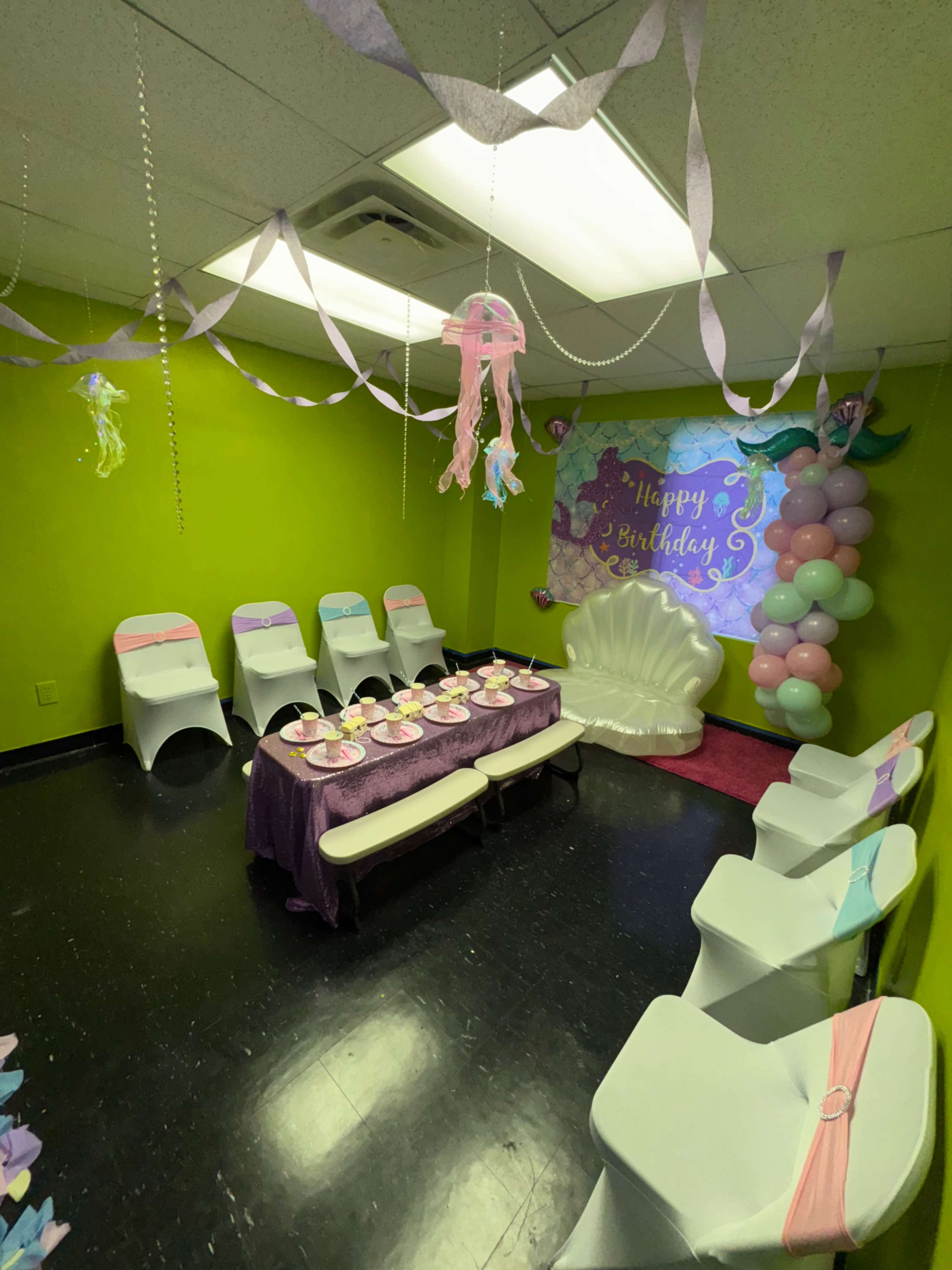 The image shows a decorated party room with green walls, a table set with birthday treats, and colorful balloons and streamers hanging from the ceiling.
