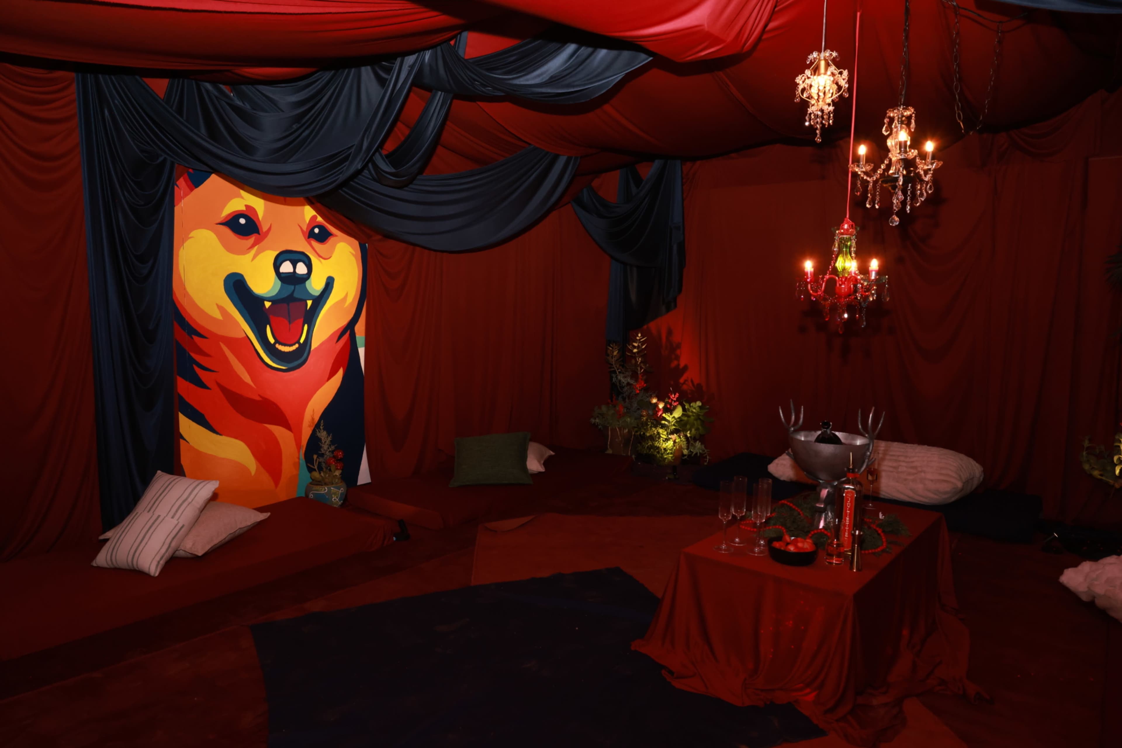 The scene depicts a cozy, dimly lit room with red drapery, a large vibrant painting of a dog on the wall, and a table set with drinks and decorative elements.