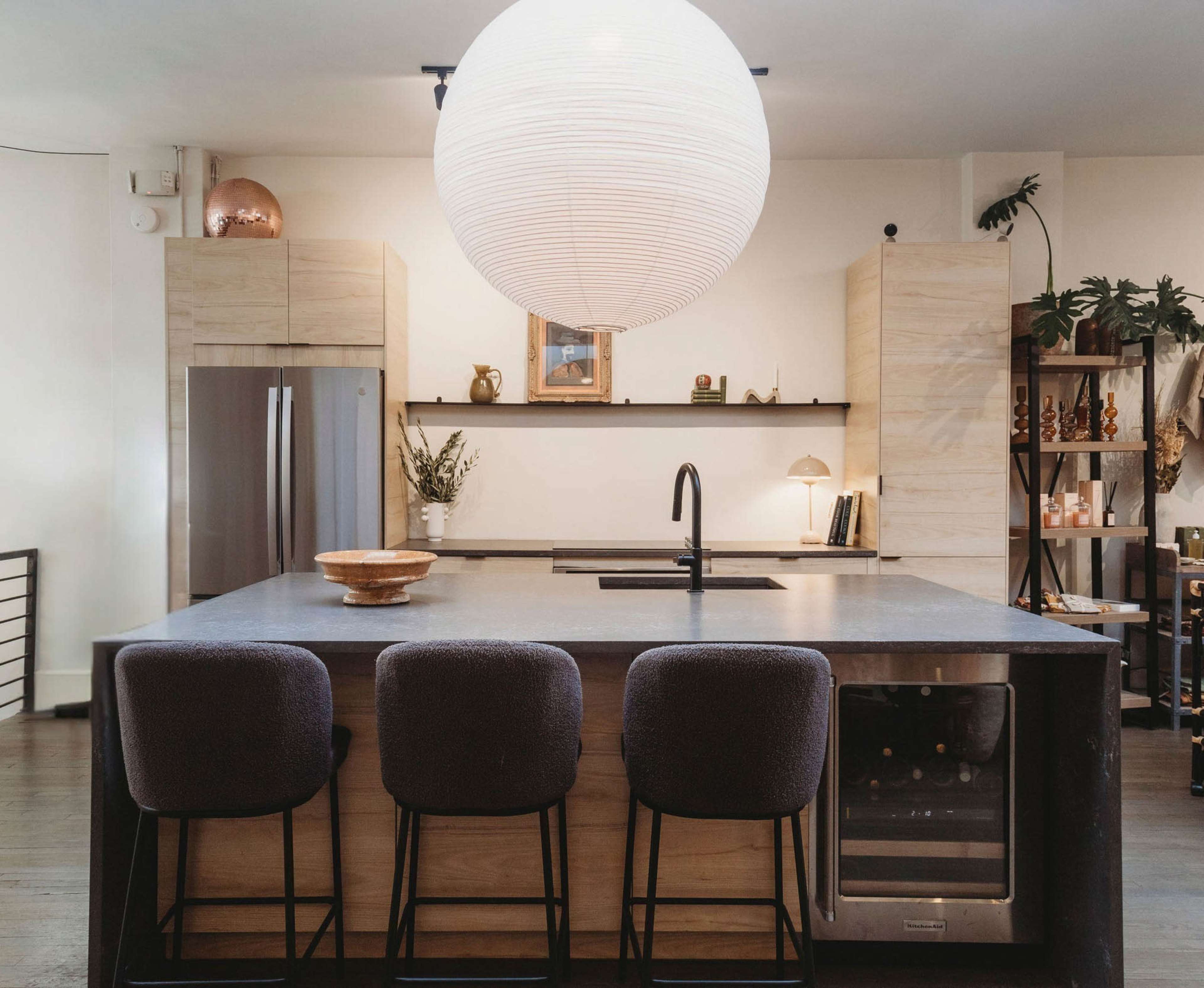 The image shows a modern kitchen with a central island, three bar stools, and a large round pendant light overhead.