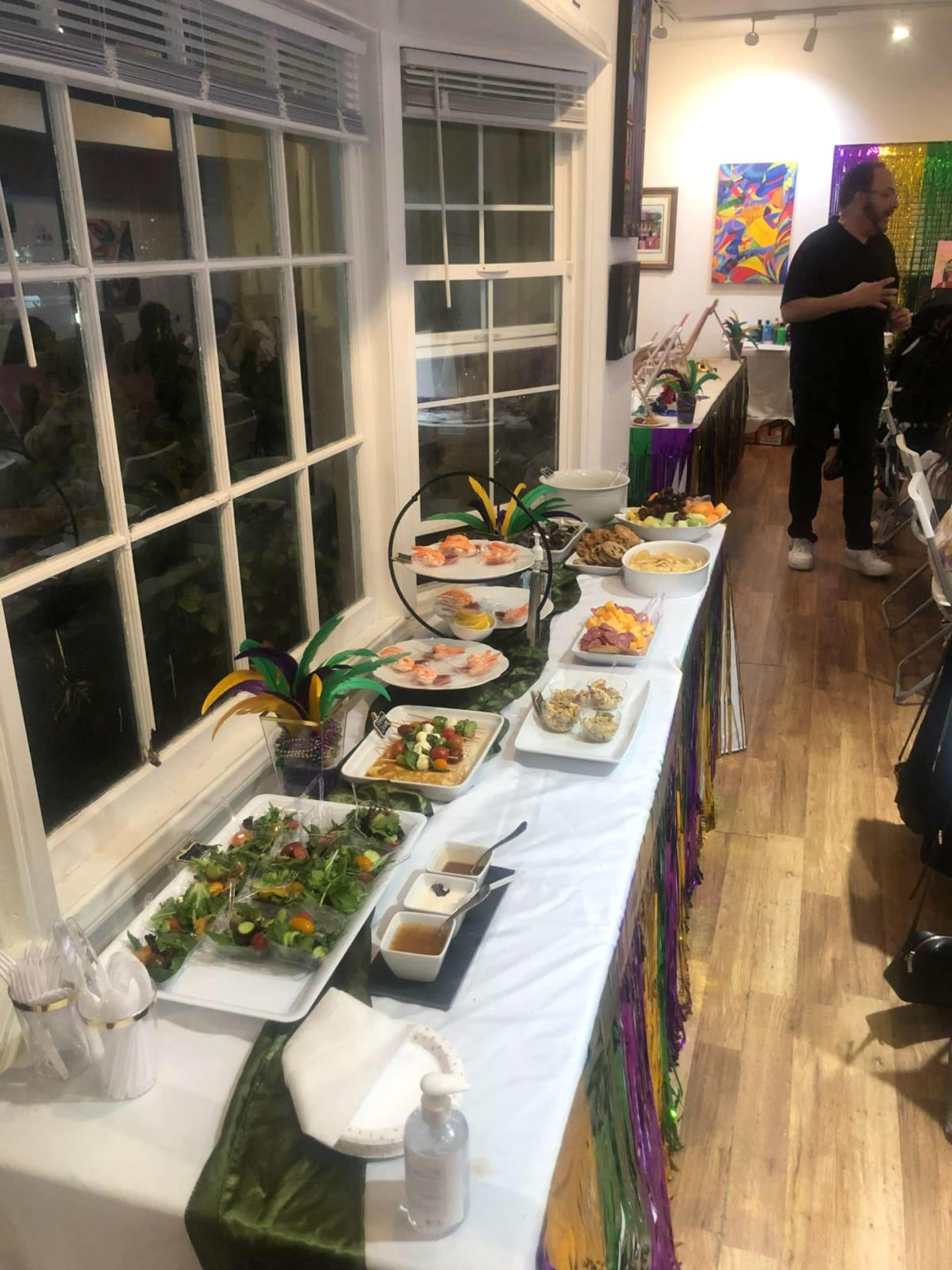 A buffet table is set up with various dishes including salads, seafood, and desserts, decorated with colorful Mardi Gras-themed accents.