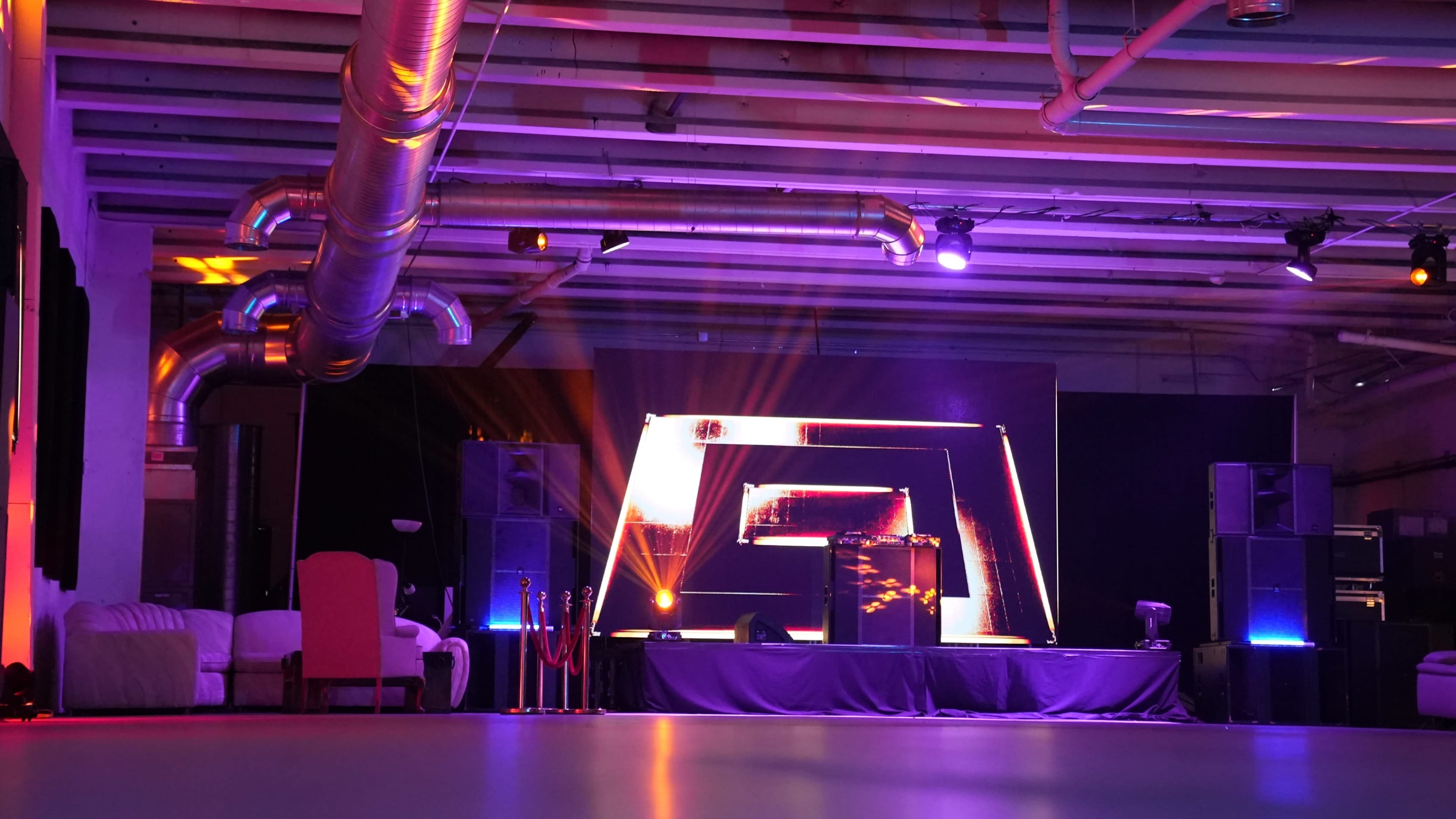 The image shows a dimly lit event space with a DJ stage featuring colorful lights, large speakers, and a row of velvet ropes in front.