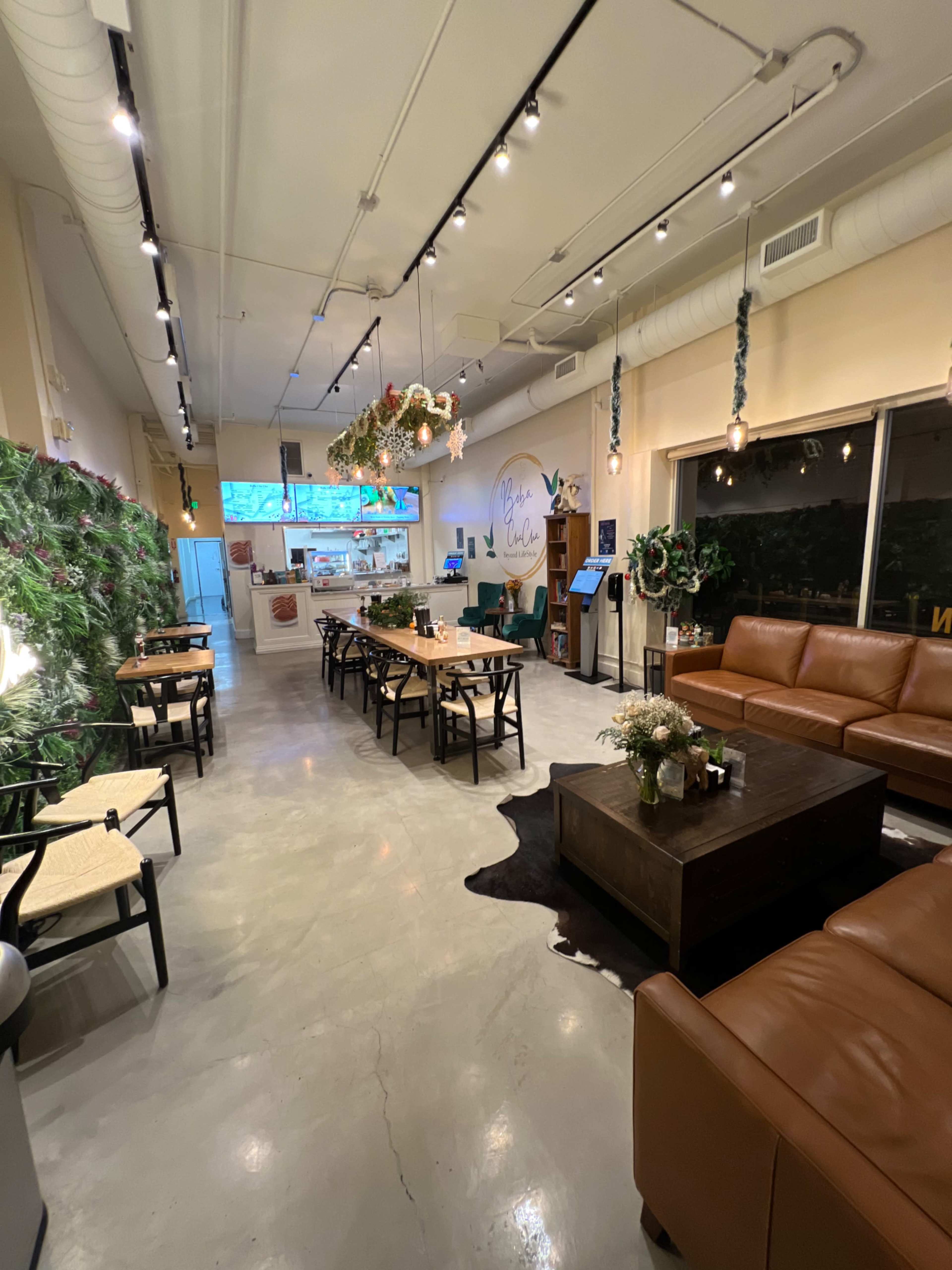 A spacious cafe interior with a mix of seating arrangements, including tables, chairs, and a couch, adorned with plants and decorative elements.