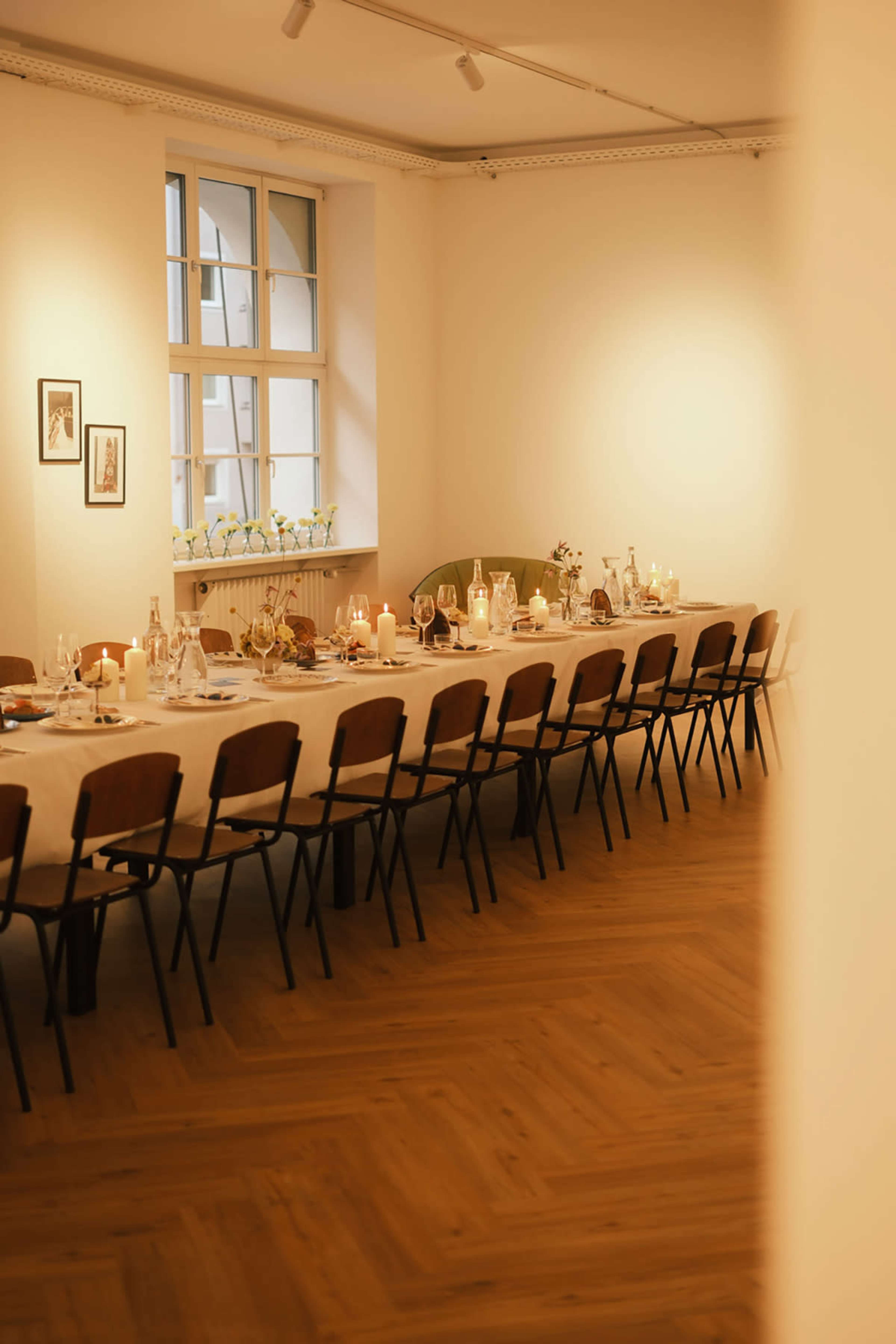 A long dining table is set with candles and tableware in a softly lit room featuring large windows.