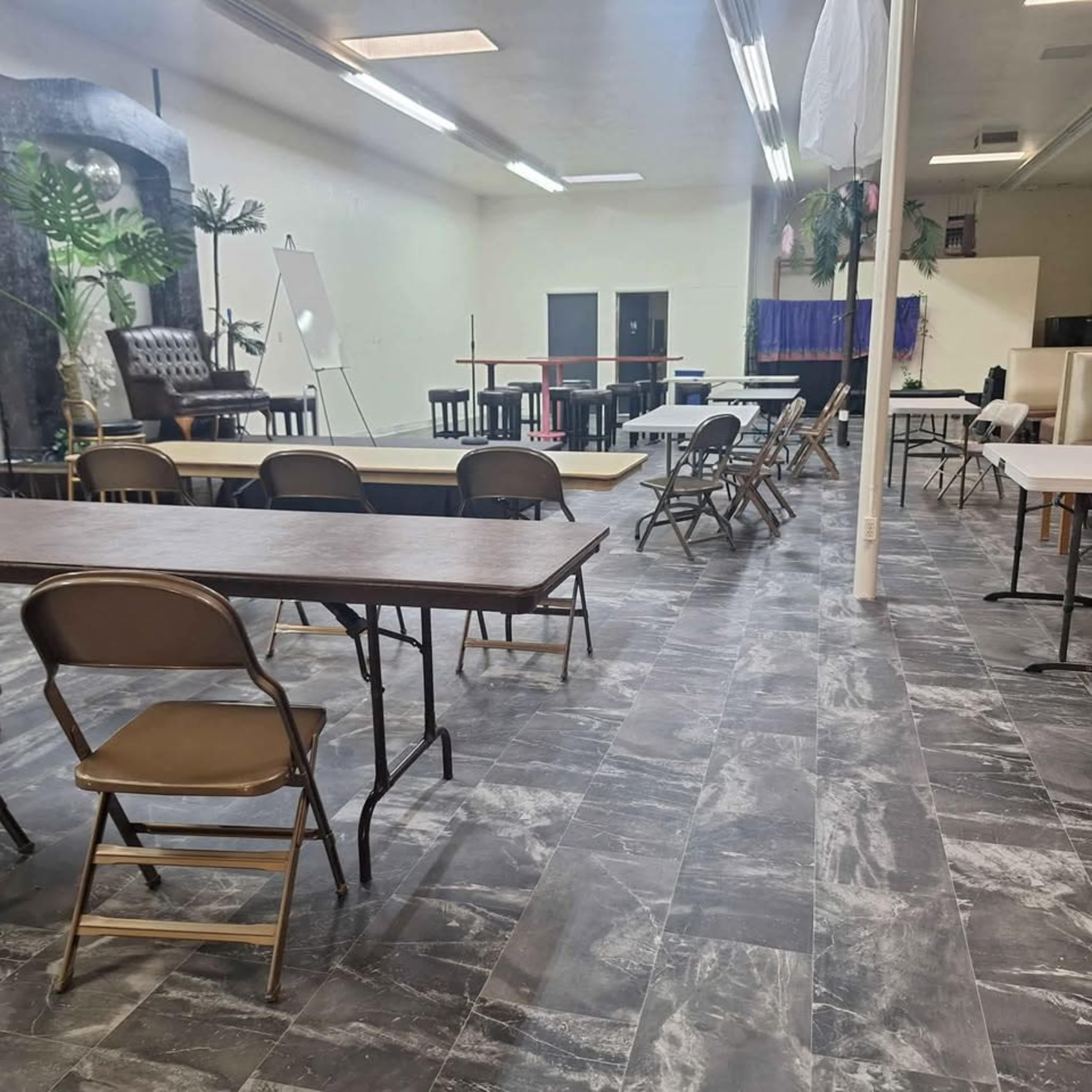 The image depicts a spacious room set up with several tables and chairs arranged for an event, featuring a stage area at one end.