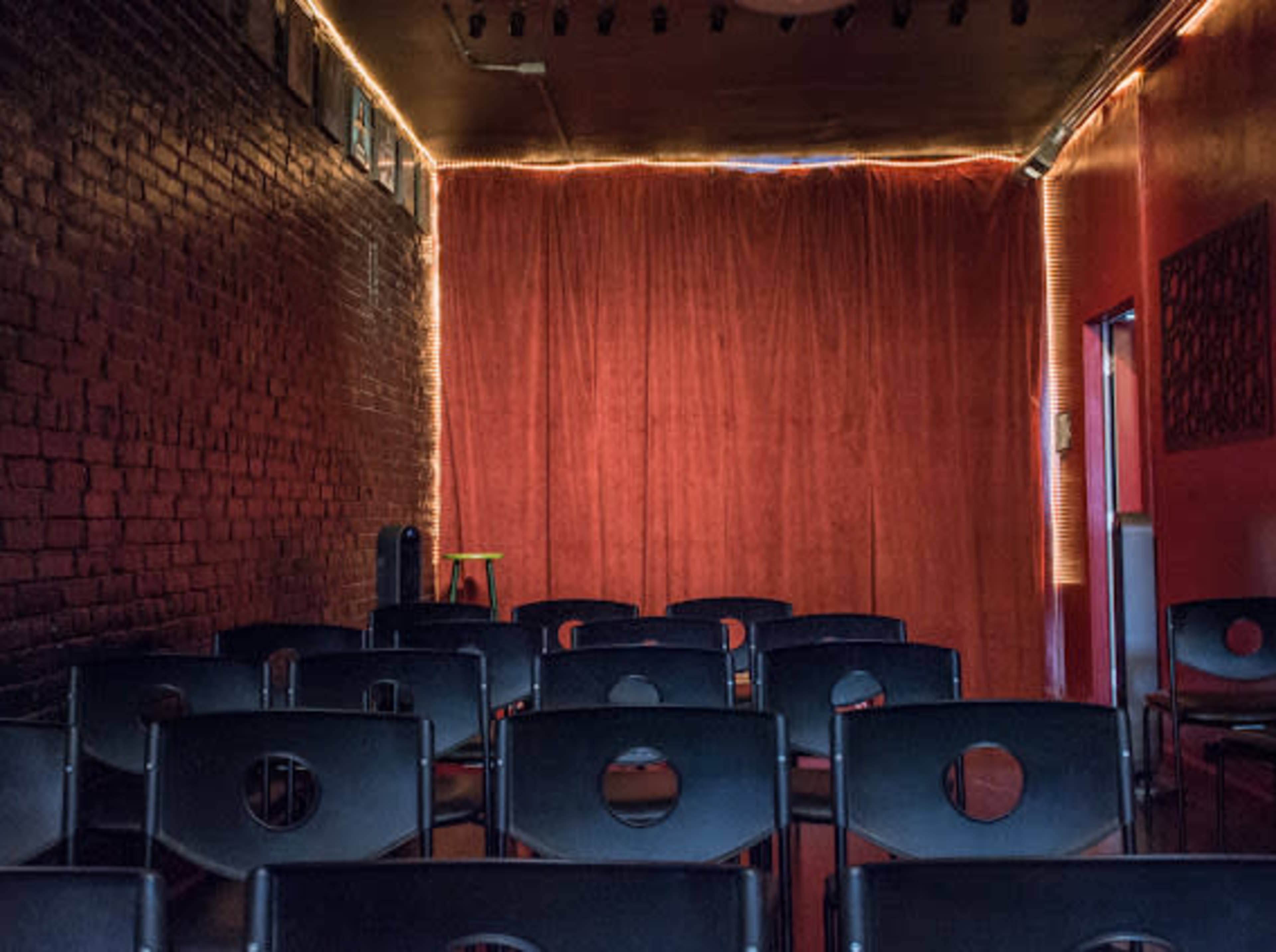 An empty performance space features rows of black chairs facing a red curtain illuminated by soft lights along the perimeter.