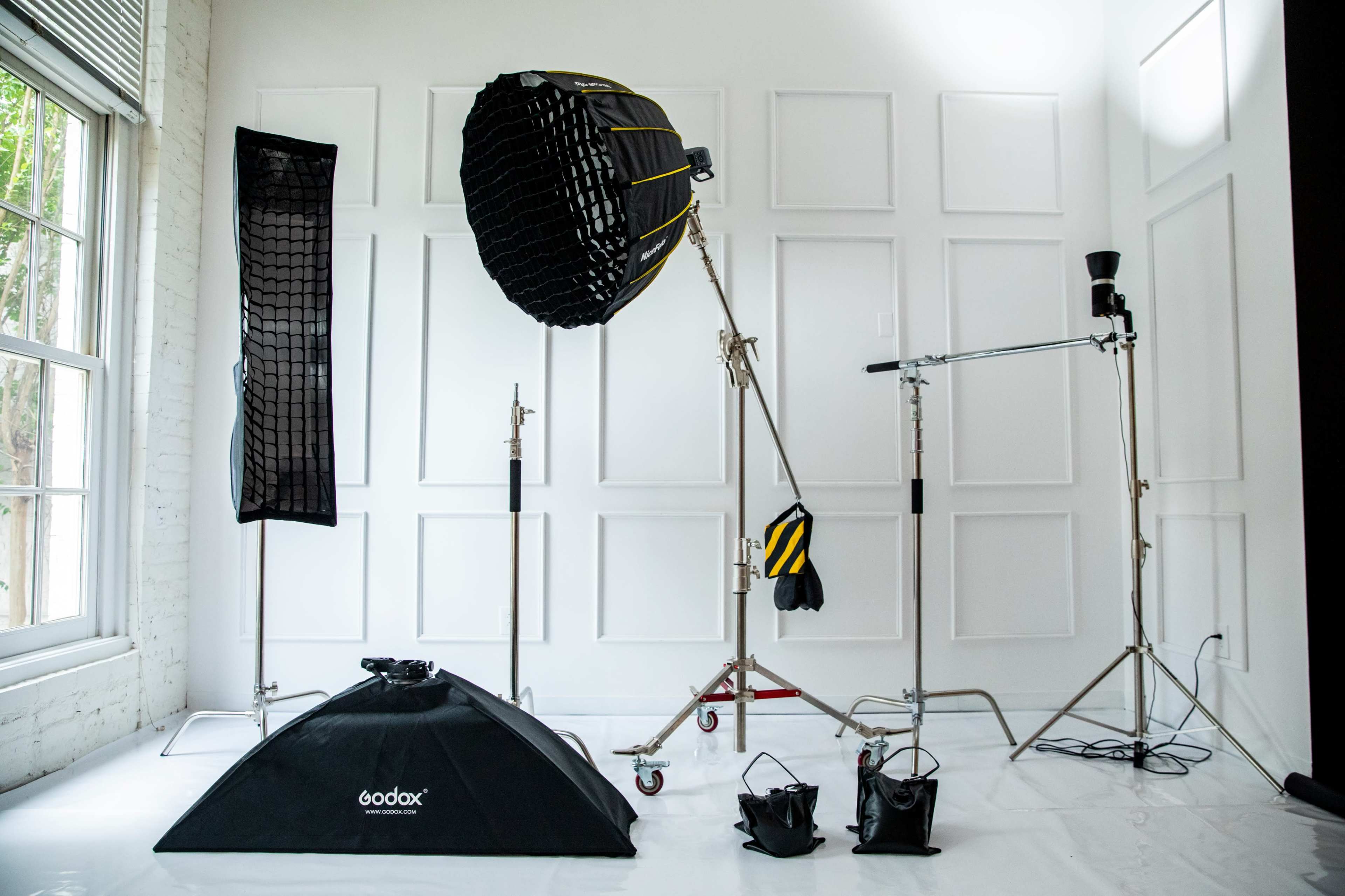 A photography studio setup featuring various lighting equipment, including a large softbox, a light stand, and backdrop supports against a white wall.