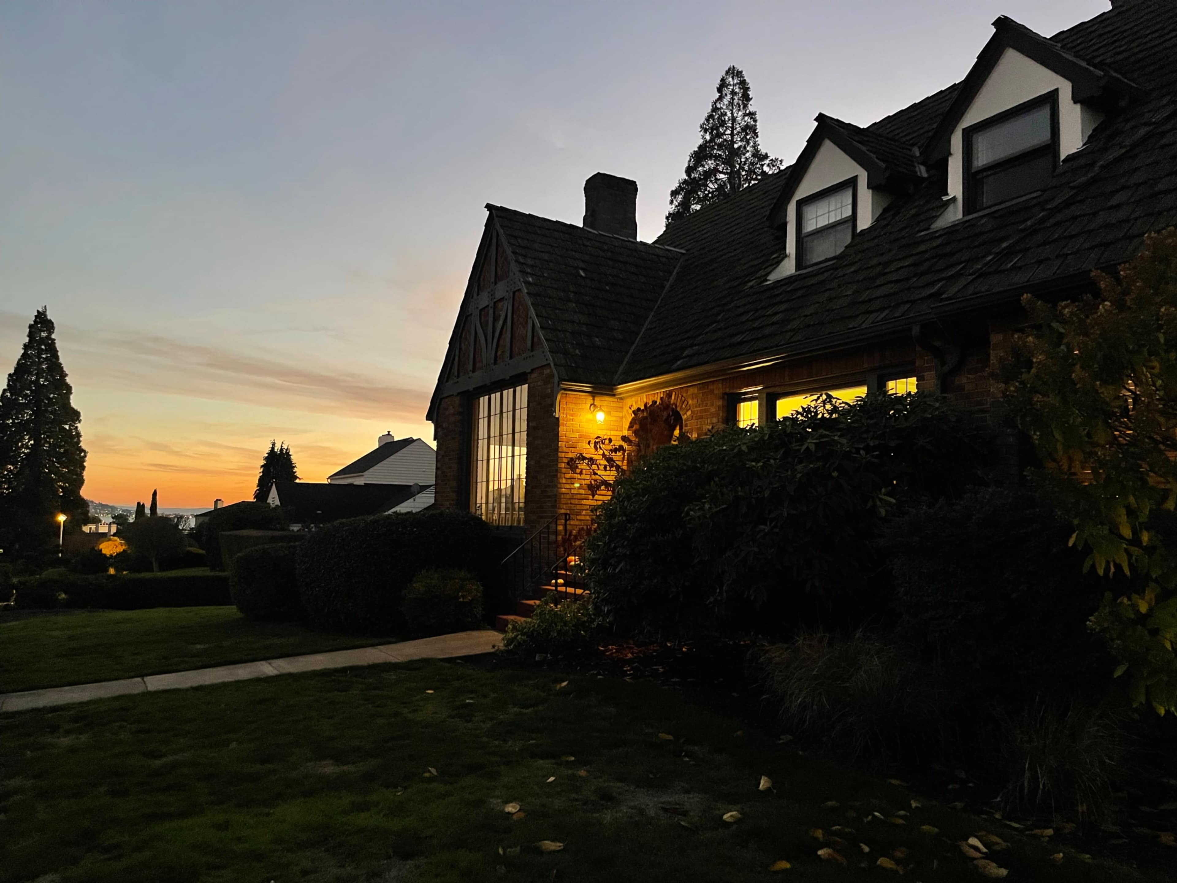 A brick house with a gabled roof is illuminated by a porch light as the sun sets in the background.