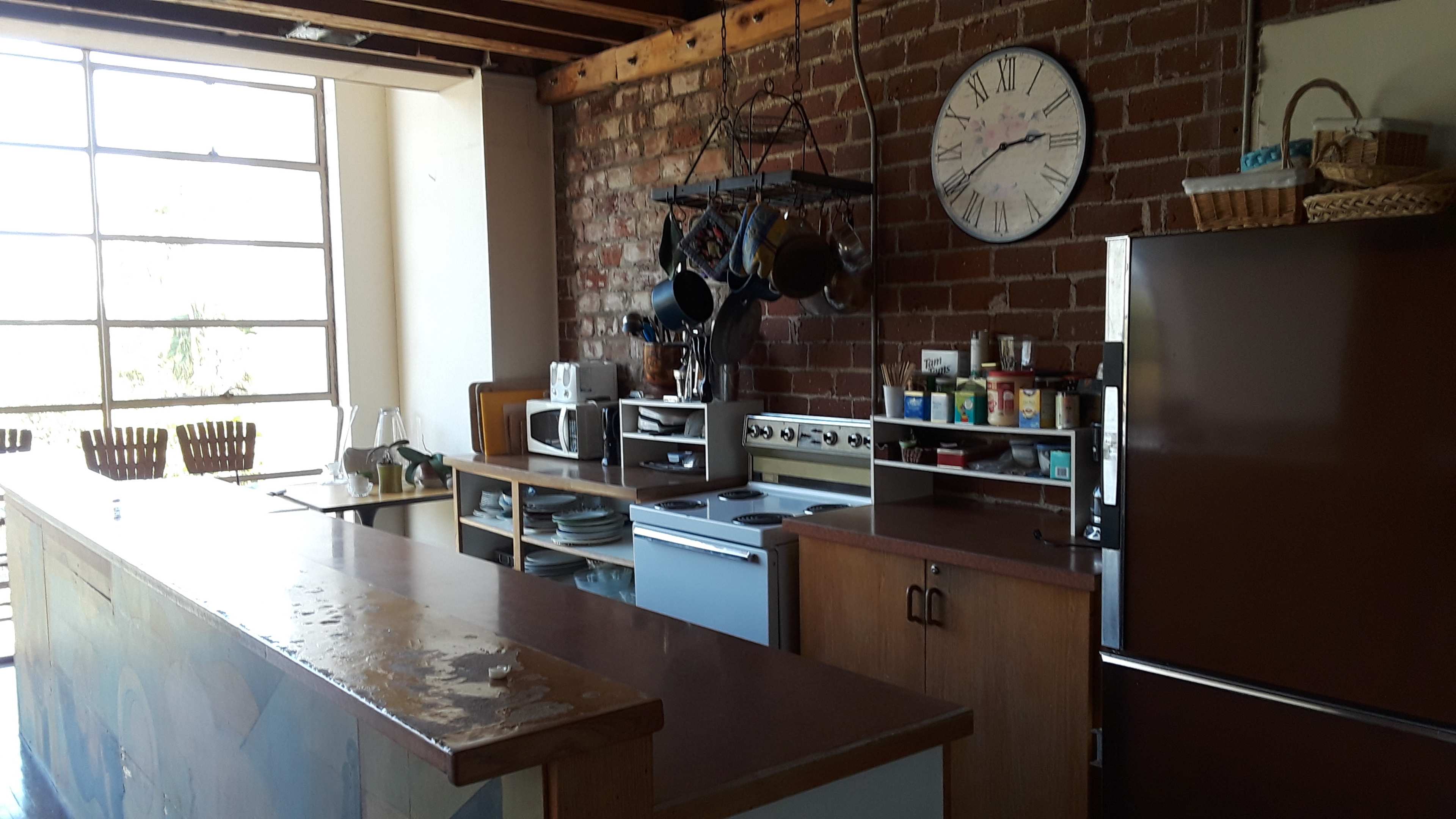 A kitchen with exposed brick walls, a large window, and various appliances including a refrigerator, stove, and microwave, along with hanging pots above the counter.
