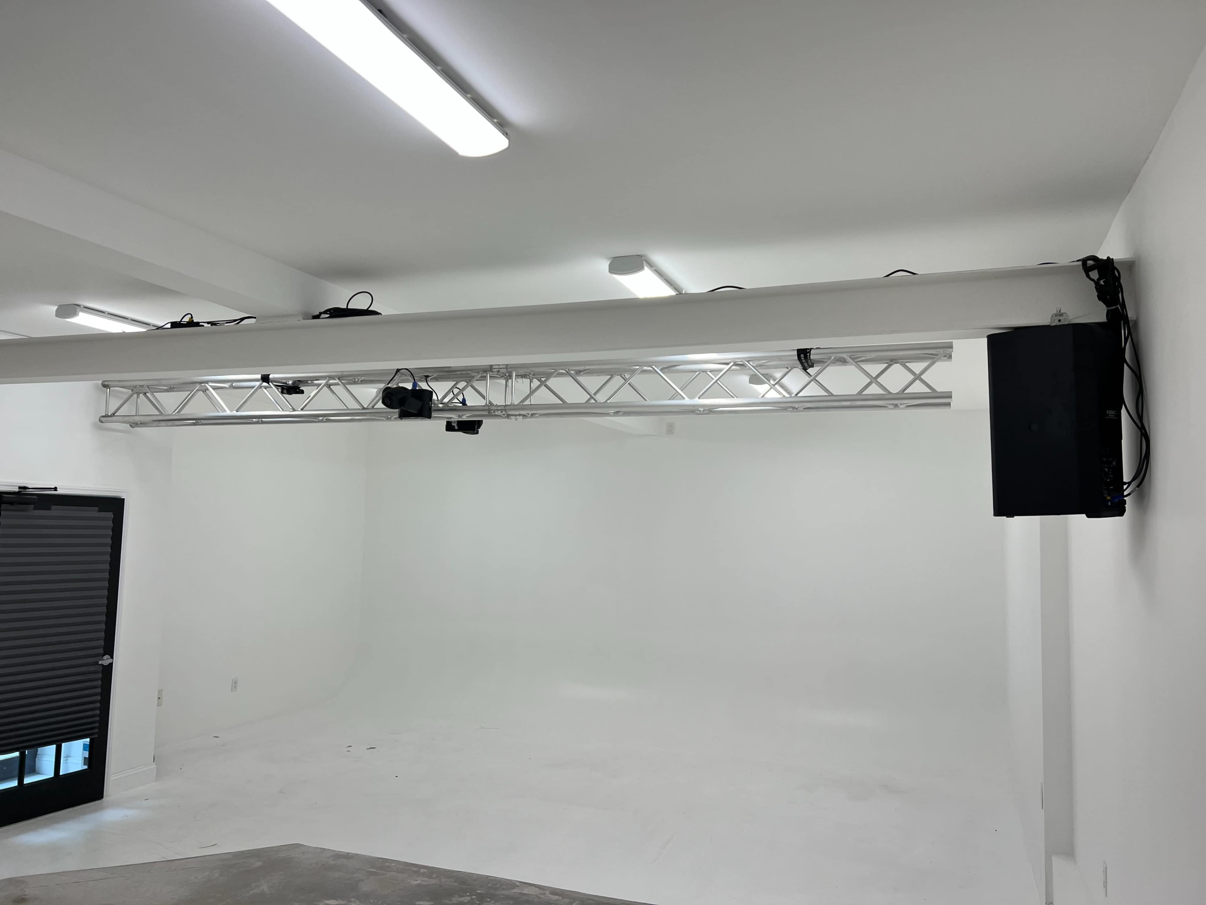 An empty studio space with a white backdrop, overhead lighting, and a mounted truss system.