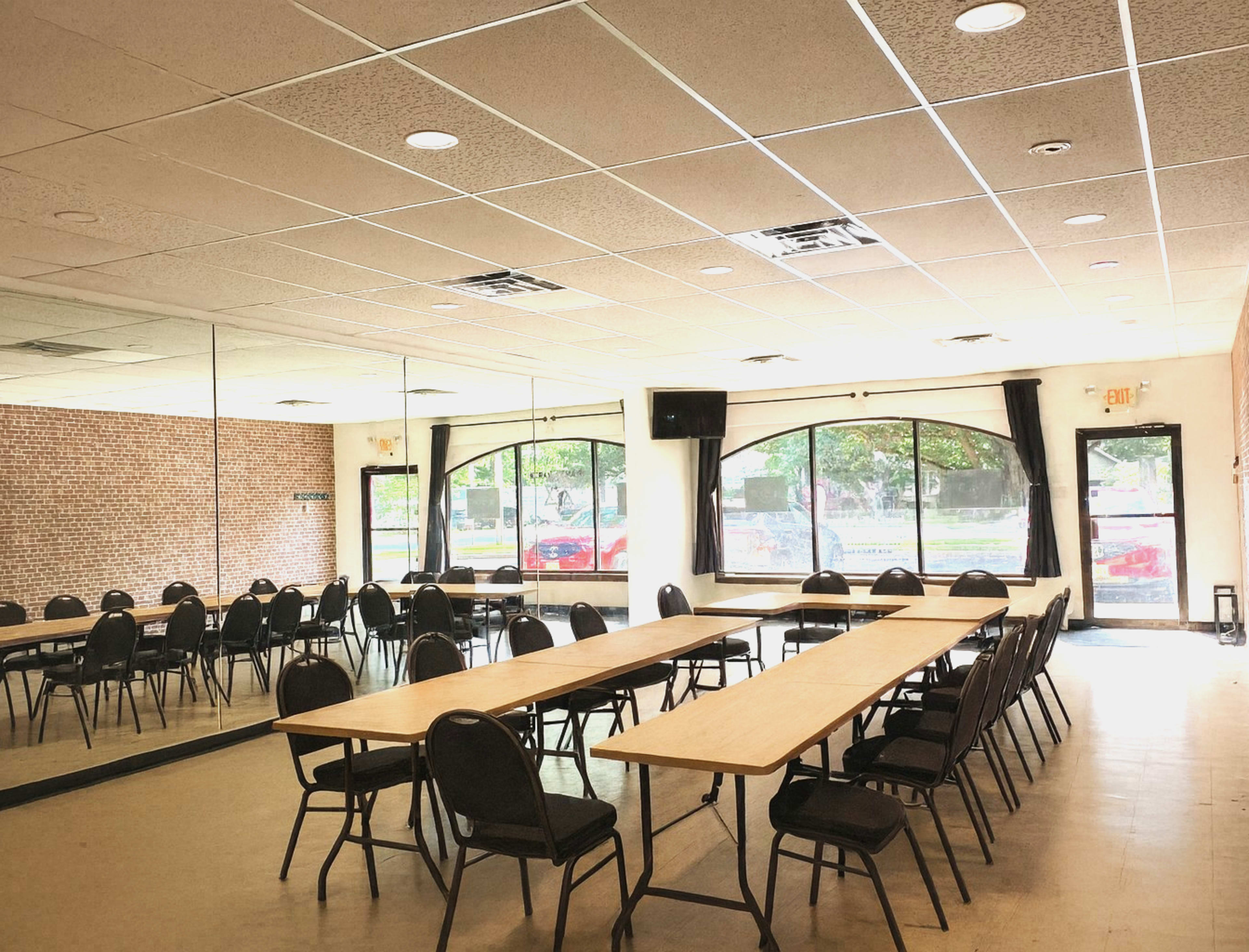 The image shows a meeting room with multiple rectangular tables arranged in a U-shape and rows of black chairs facing them, against a backdrop of large windows and a mirrored wall.