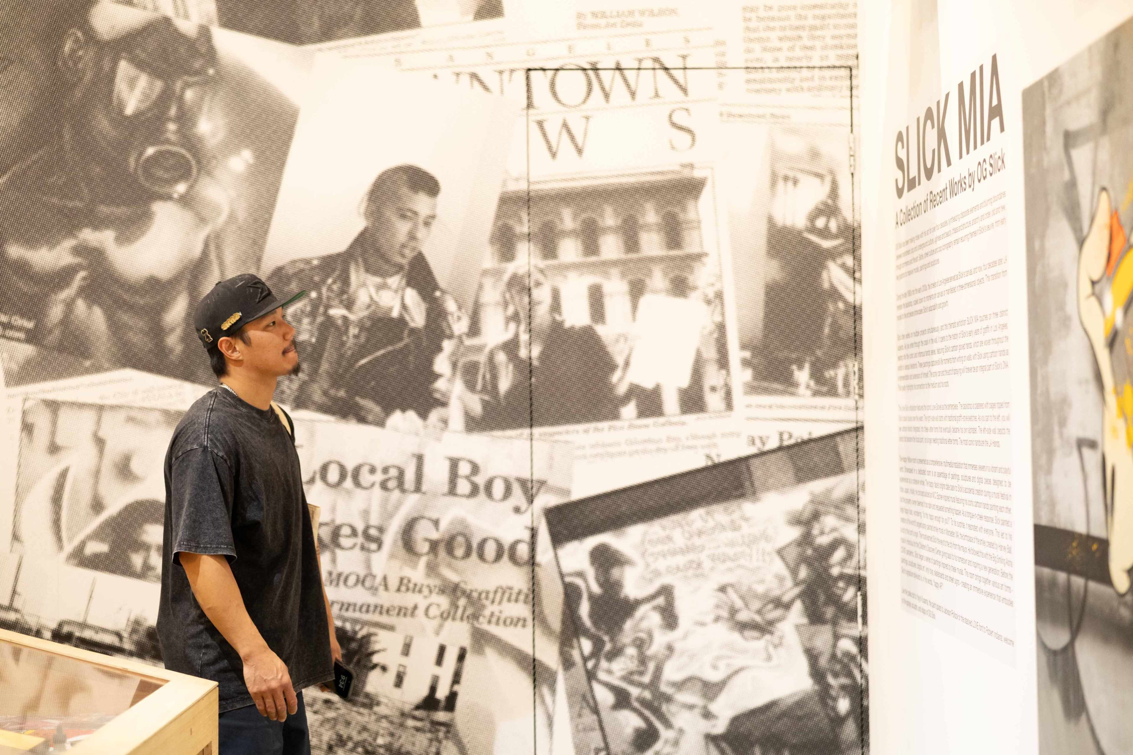A man stands in an art exhibit, looking at a wall display titled "SLICK MIA" surrounded by black and white photographs and newspaper clippings.