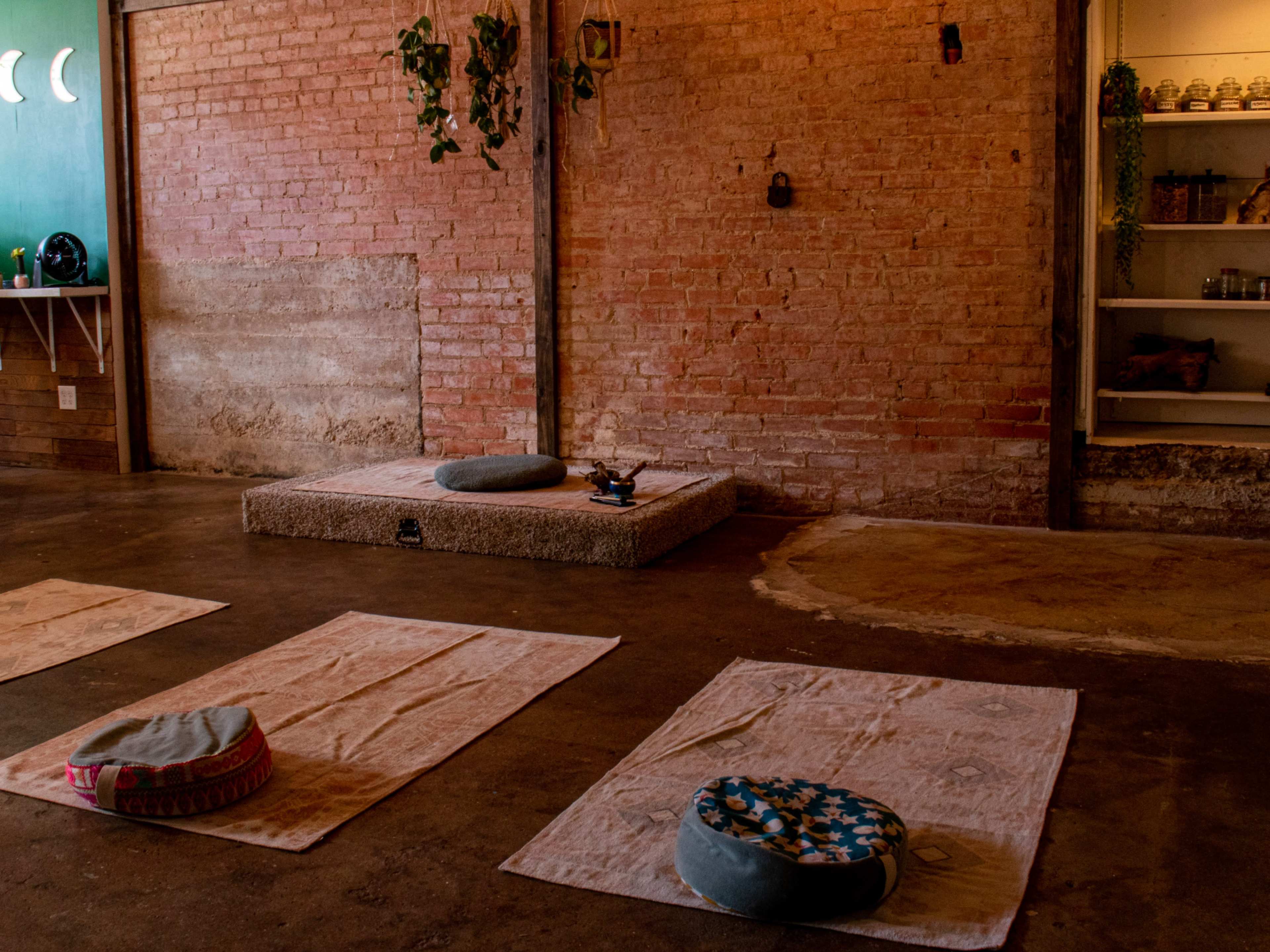 The image shows a serene yoga studio with a wooden floor, several mats laid out, and a raised platform containing cushions and decorative items against a brick wall.