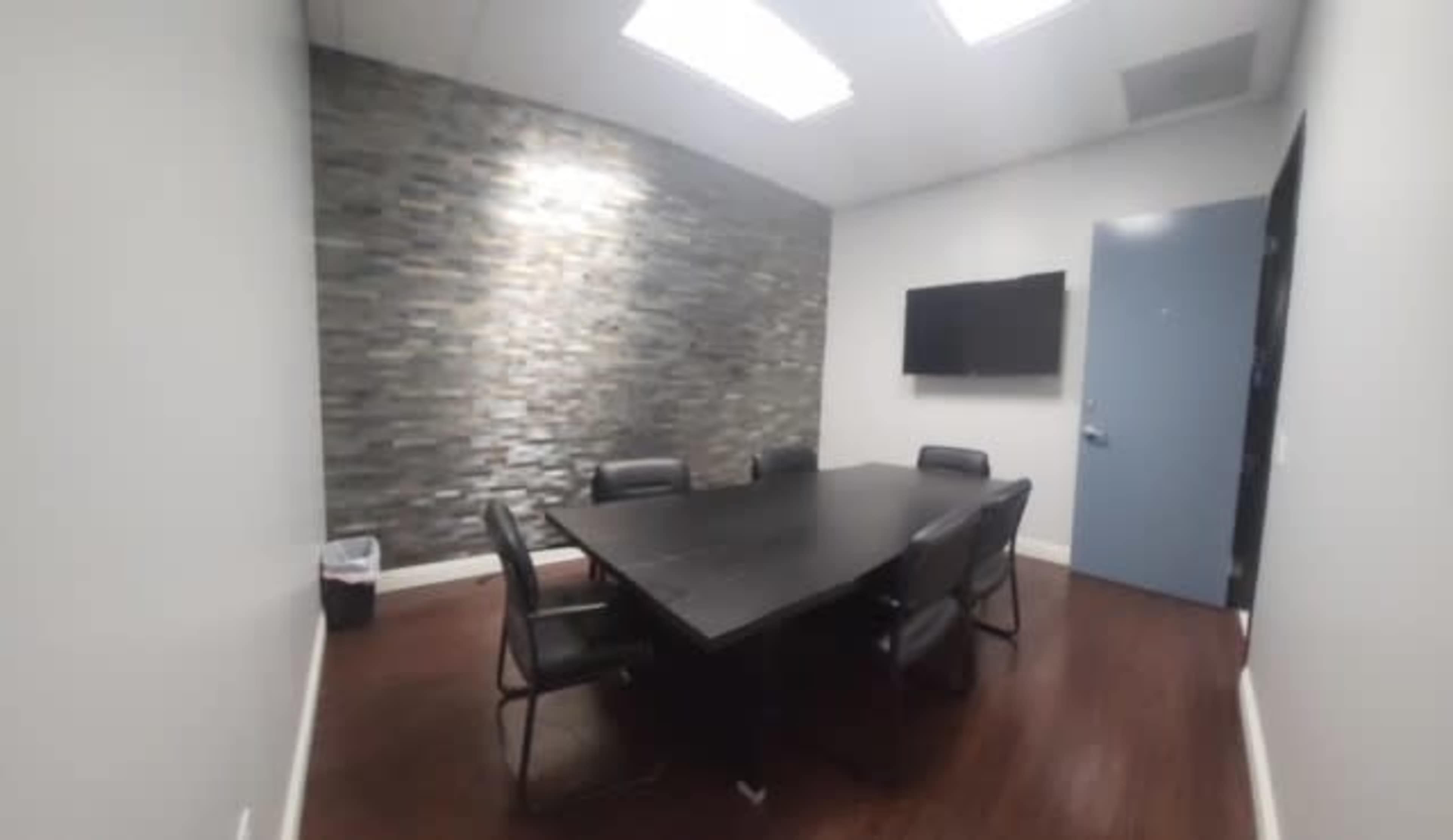 A conference room features a large black table surrounded by chairs, a stone accent wall, and a wall-mounted television.