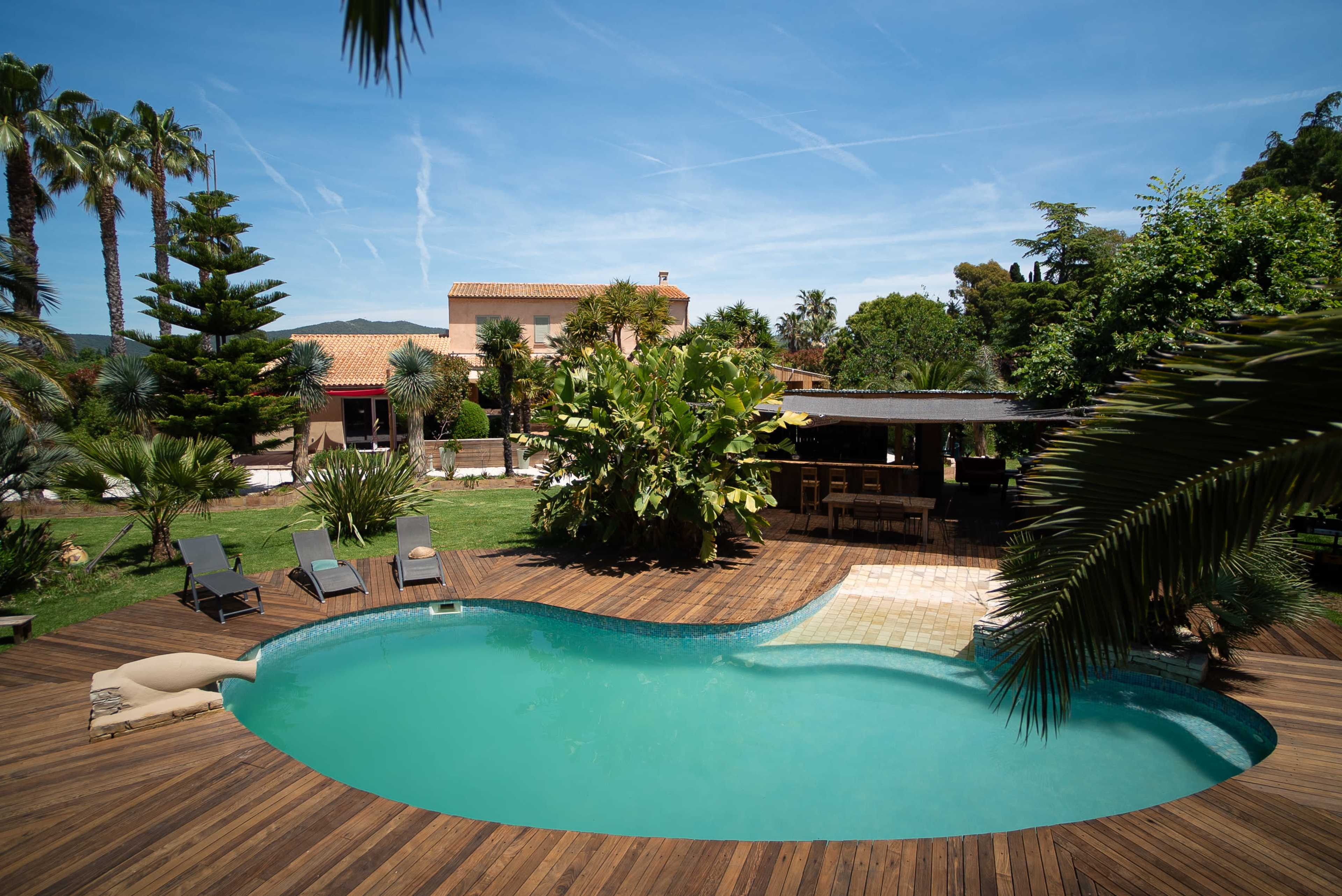 The image shows a large outdoor pool surrounded by wooden decking and lounge chairs, set in a lush garden with palm trees and a two-story house in the background.