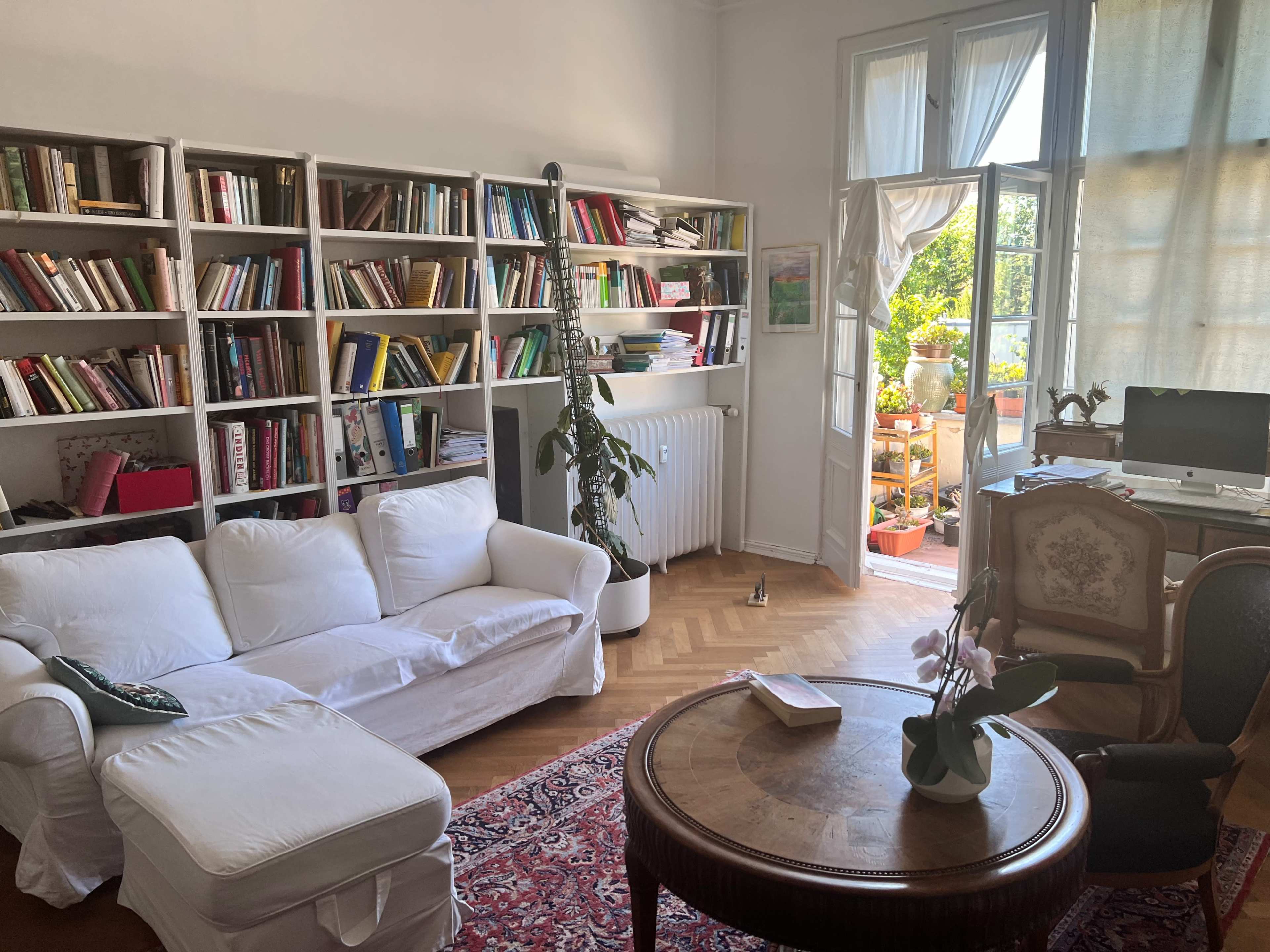 A cozy living room features a white sofa, a round wooden table, bookshelves filled with books, and a bright view of a balcony with plants.