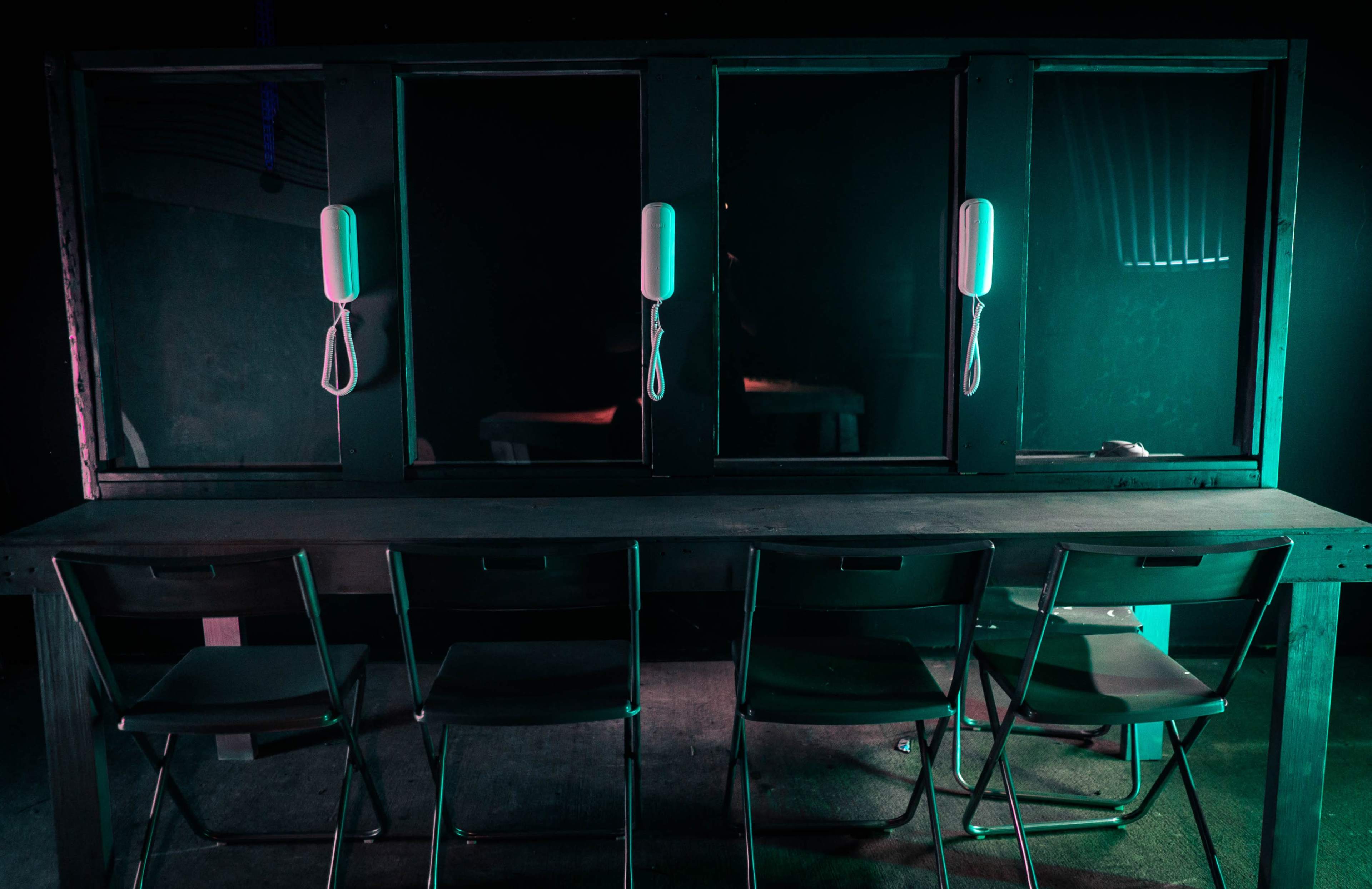 A dimly lit room features a long table with four metal chairs facing a glass partition containing two hanging phone receivers.