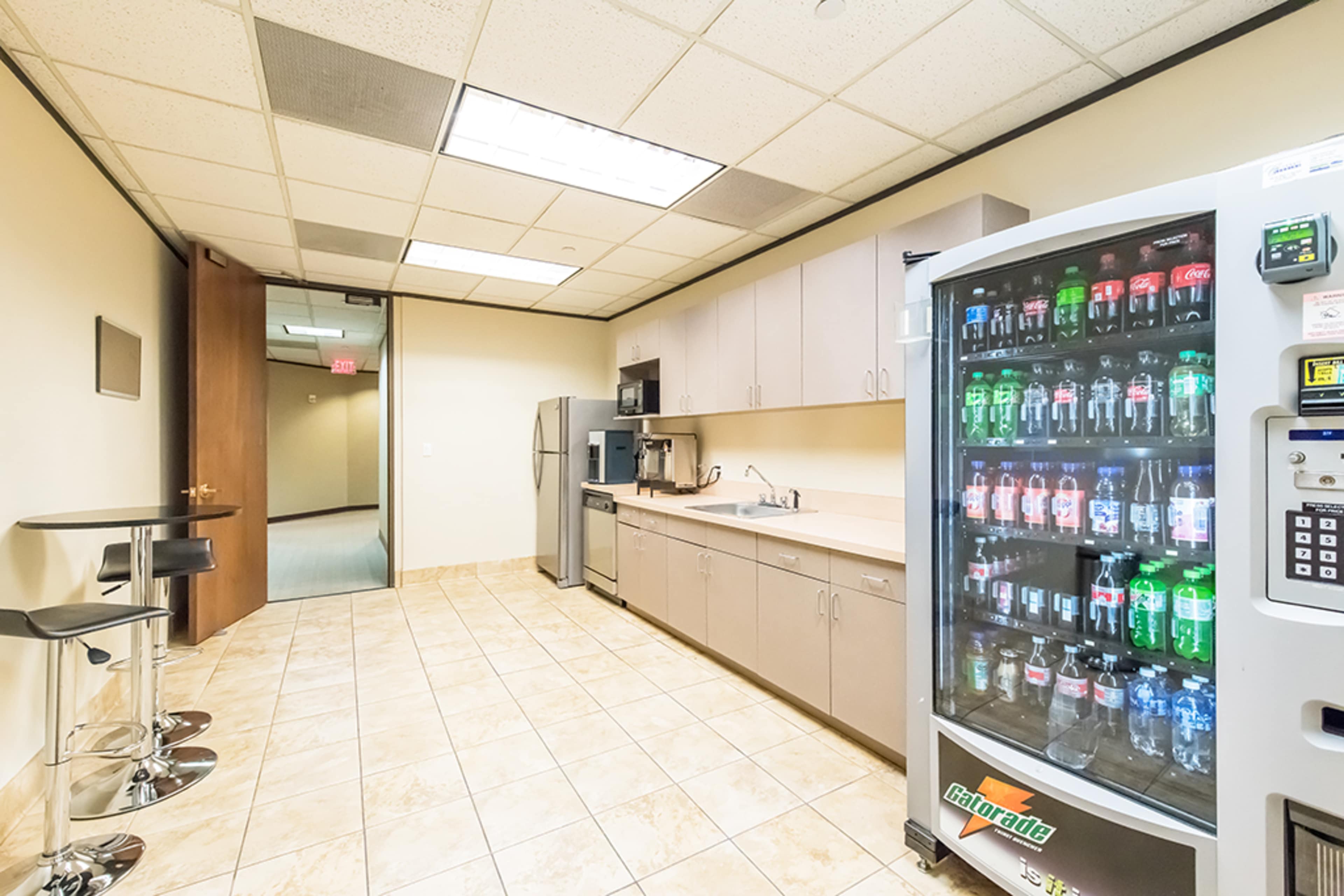 The image shows a break room with a kitchenette featuring a refrigerator, microwave, and a vending machine stocked with various beverages, set against tiled flooring and light-colored walls.