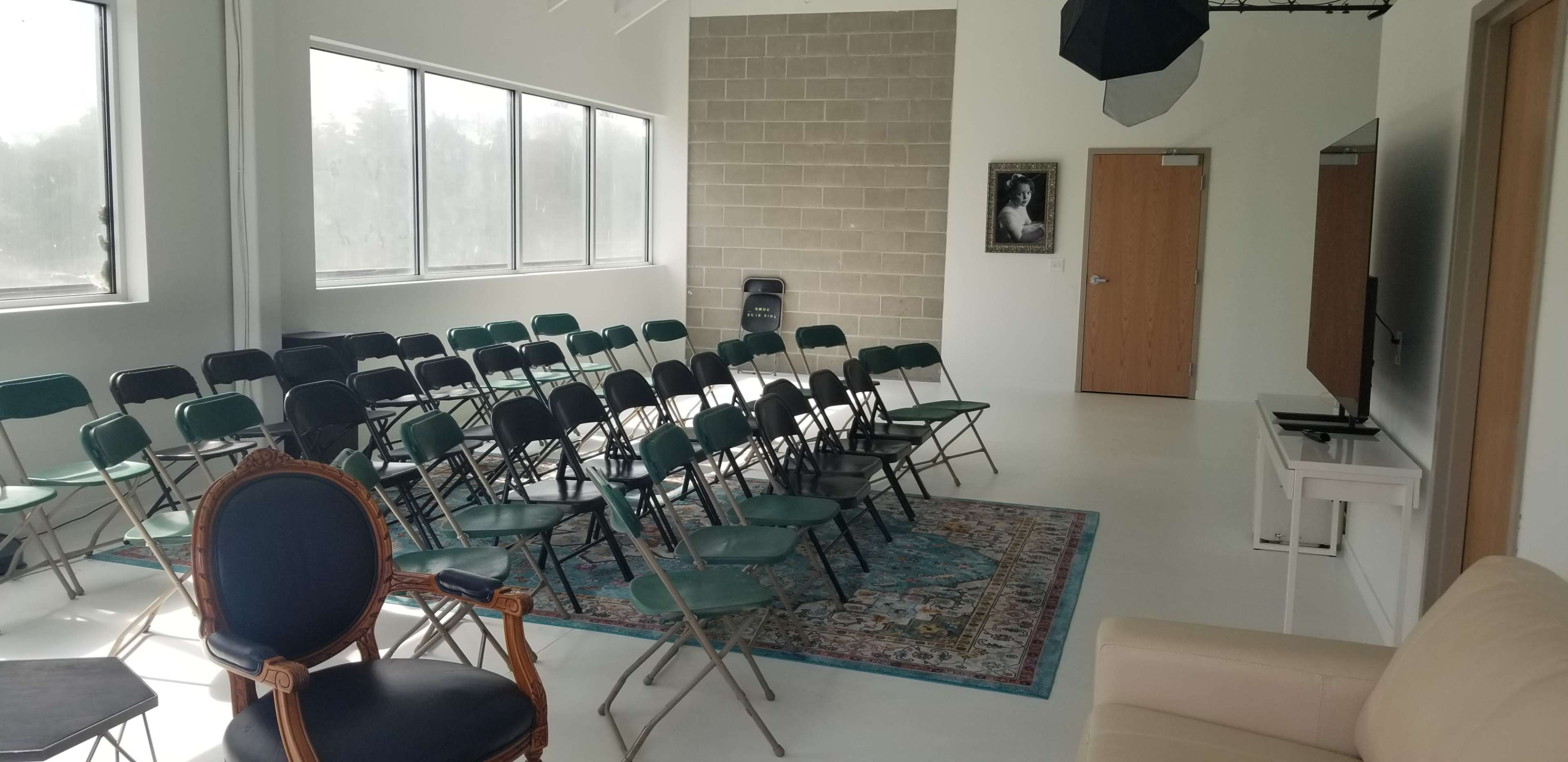 The image shows a room set up with rows of green folding chairs, a large TV on a stand, and a rug, alongside a sofa and a wooden armchair.