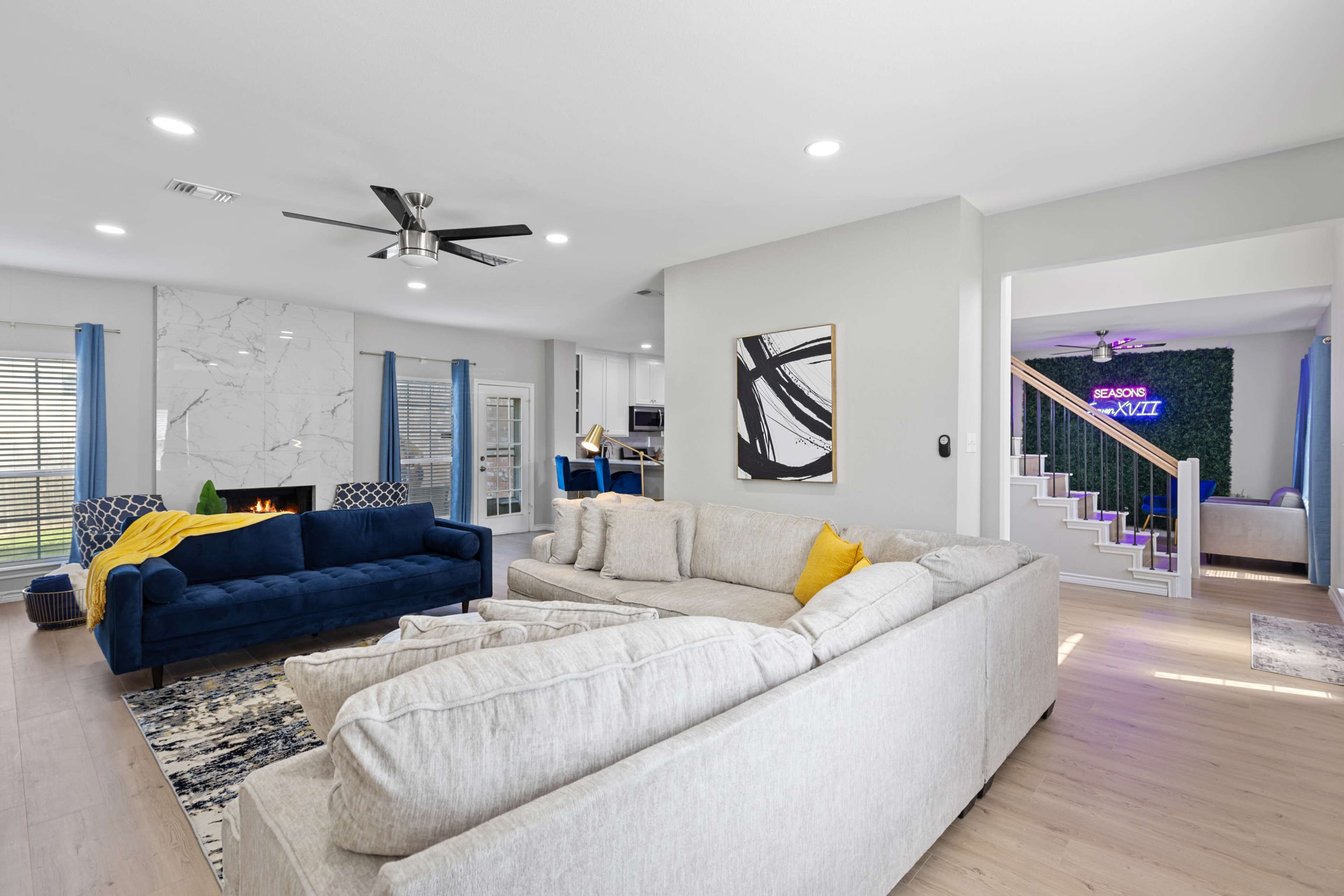 The image shows a modern living room with a blue sofa and a light-colored sectional, set against a backdrop of a marble accent wall and a staircase, with a planted space visible in the background.