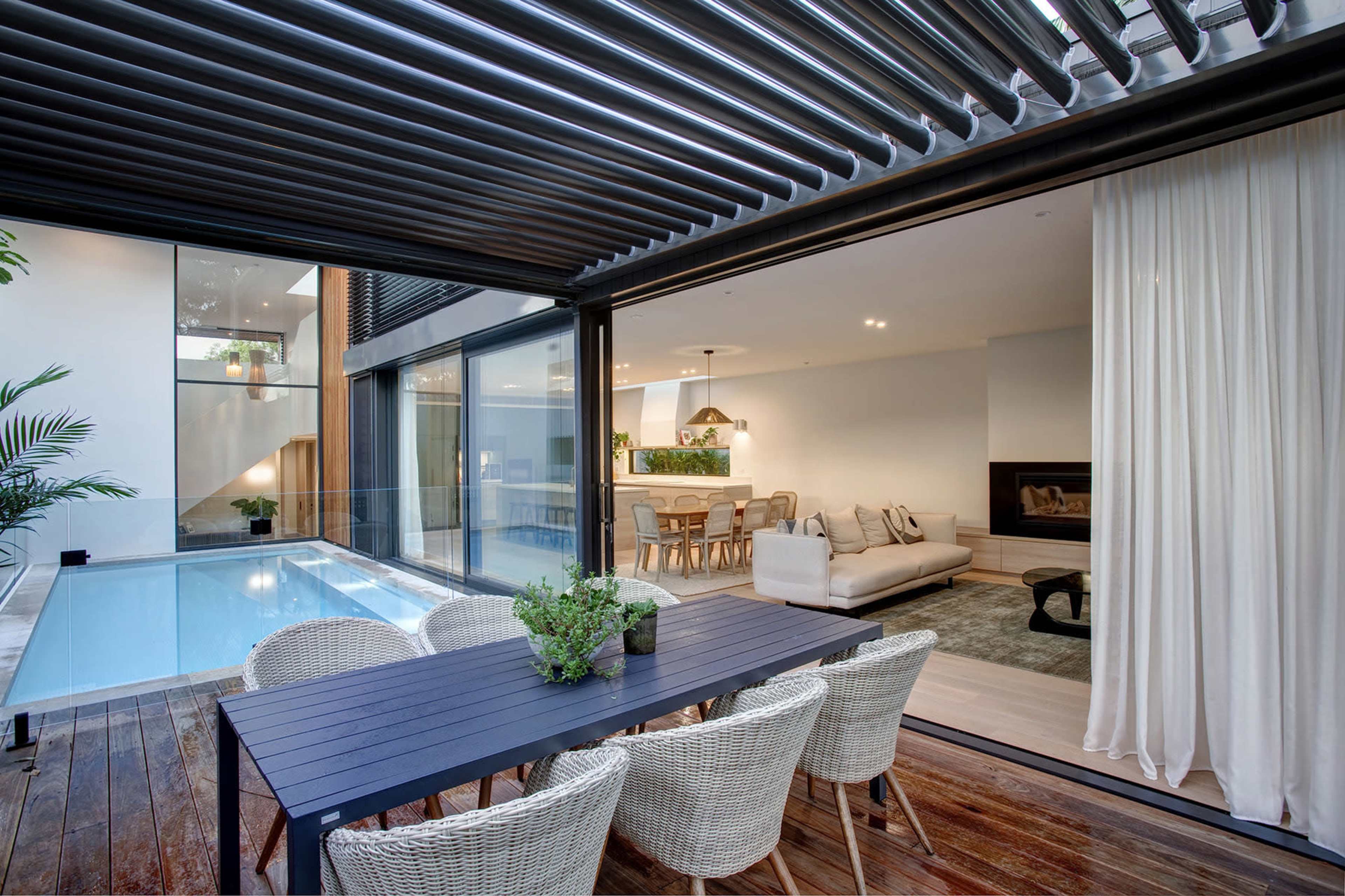 A modern indoor-outdoor living space featuring a dining area with a table and chairs next to a swimming pool, with large glass walls that open into a stylish living room.