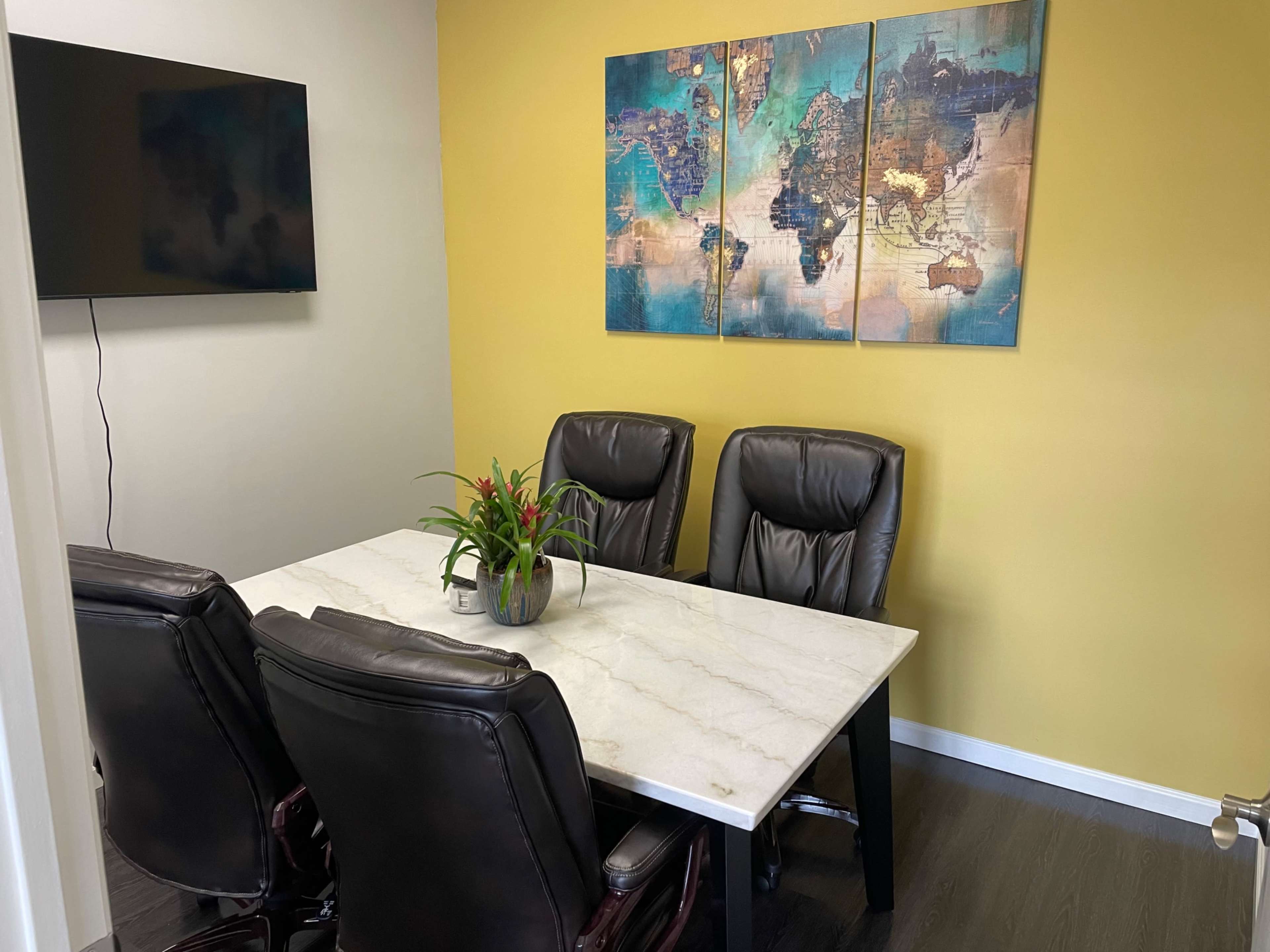 A conference room features a marble table surrounded by four black leather chairs, with a television mounted on the wall and a colorful world map artwork.
