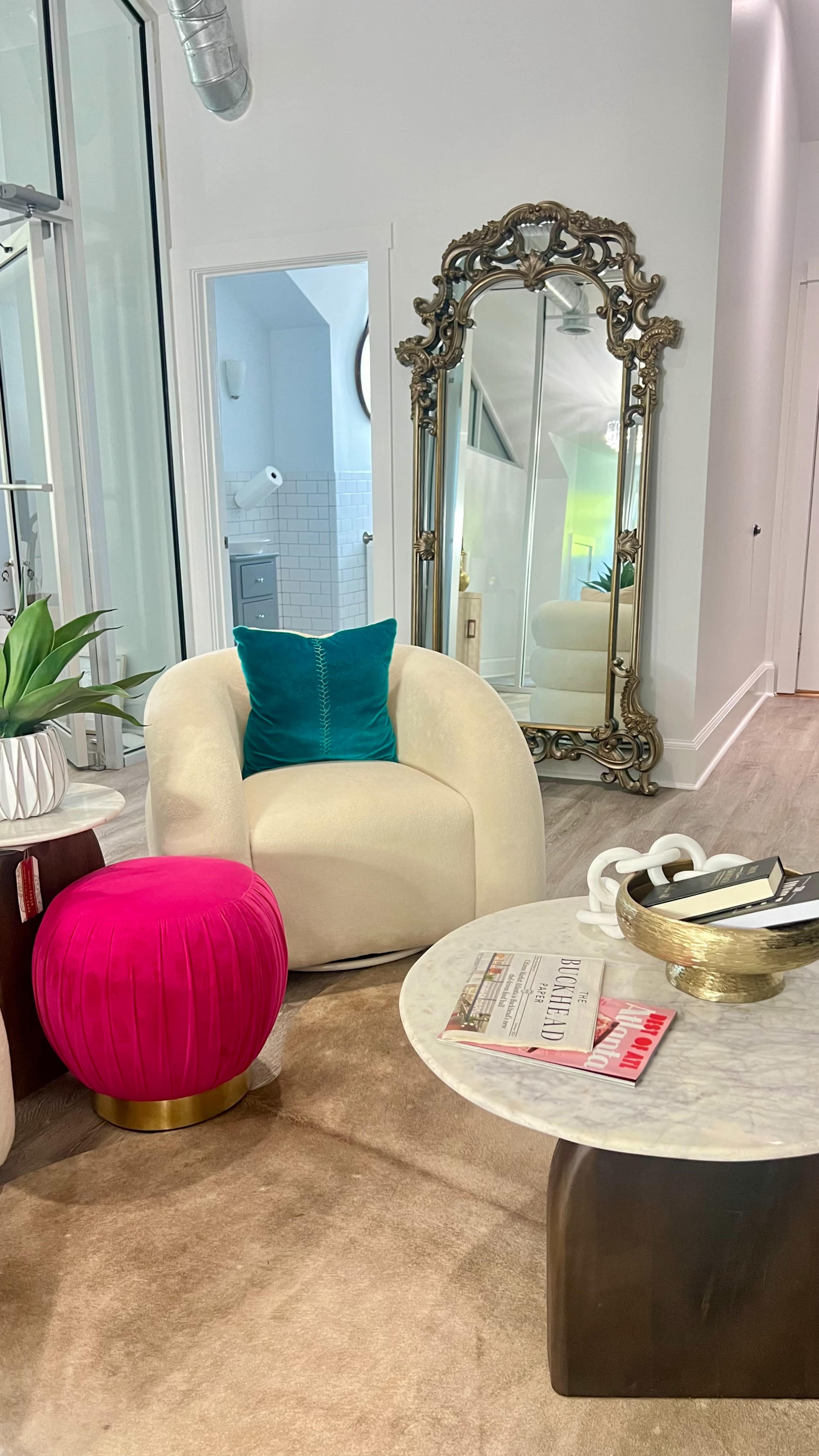 The image shows a modern living space featuring a white armchair, a bright pink pouf, a round coffee table, and a large ornate mirror against a light-colored wall.
