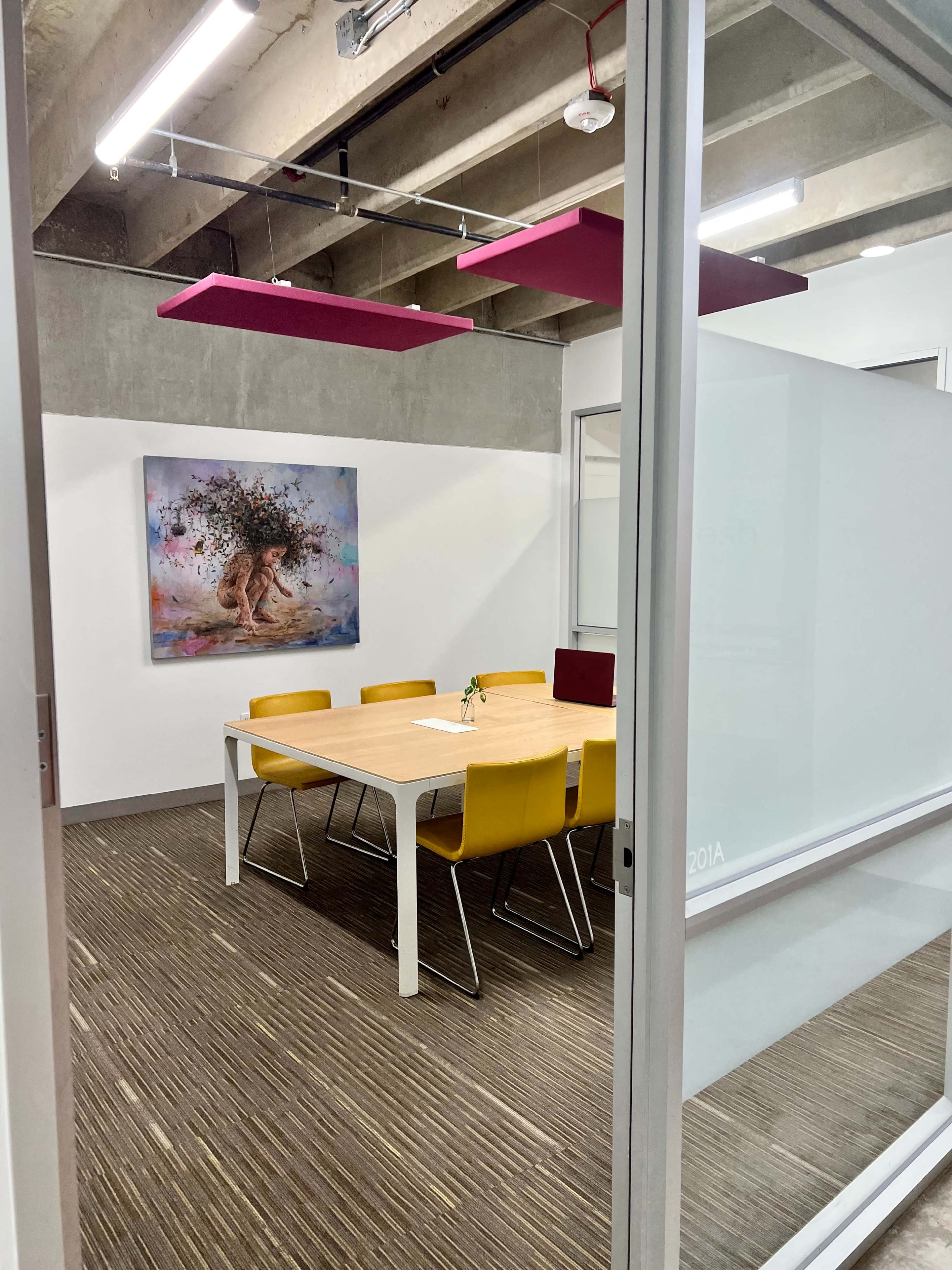 A meeting room with a wooden table surrounded by yellow chairs, a floral artwork on the wall, and pink ceiling panels.