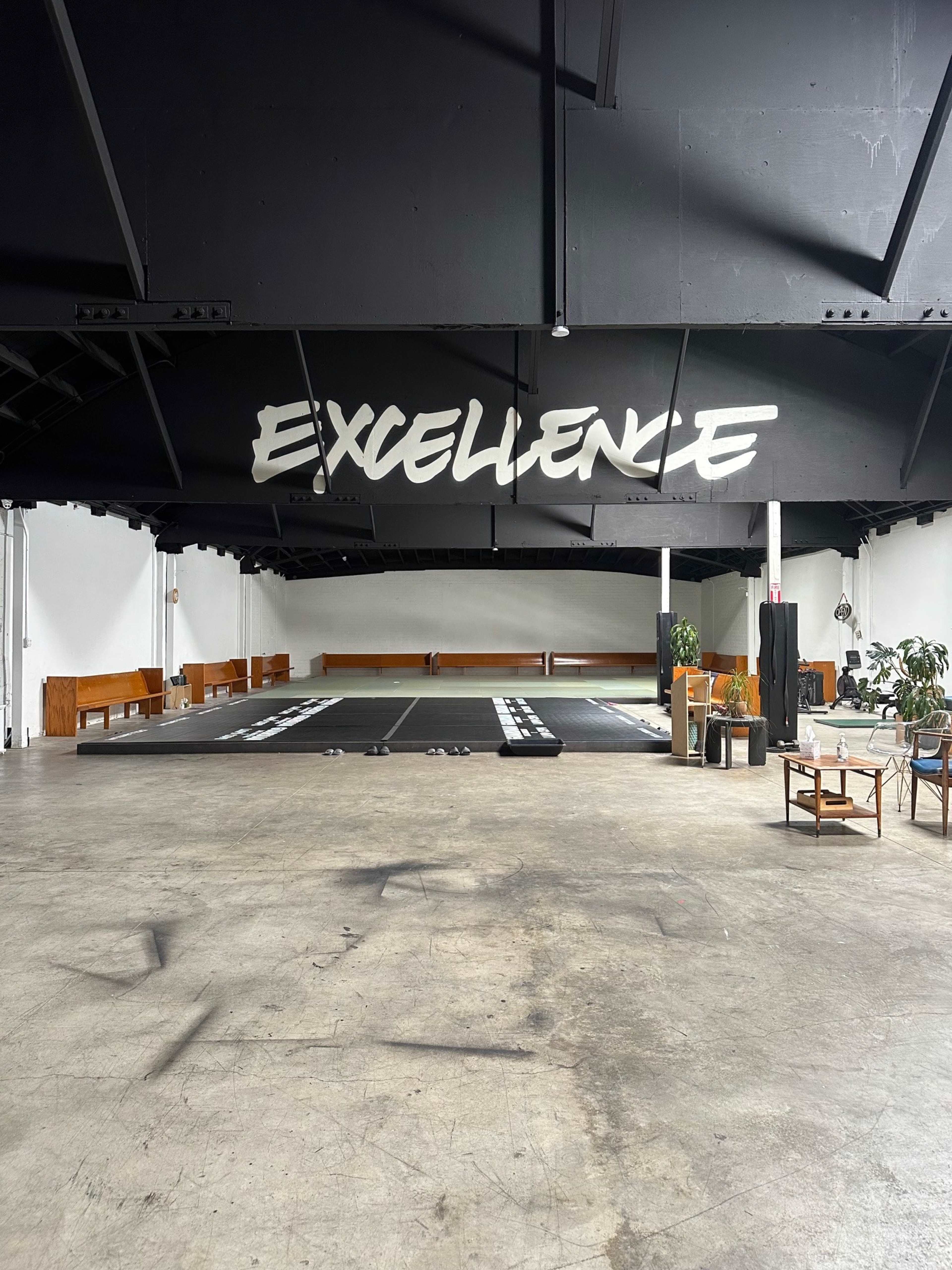The interior of a large, open space features a black ceiling with the word "EXCELLENCE" painted in white, and a floor covered in dark mats.